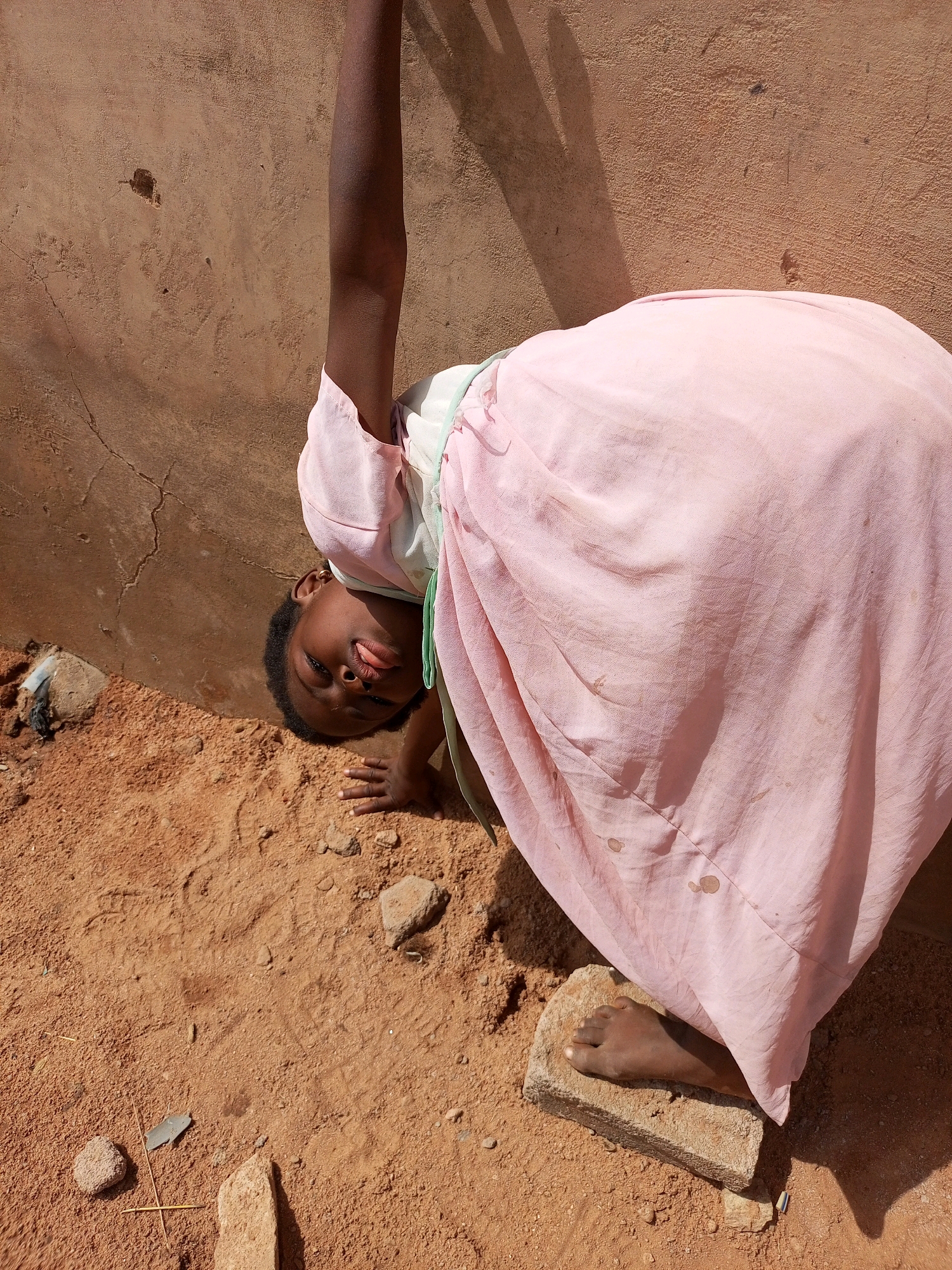 A young Black girl, seen from a tilted perspective and upside down, is in a contorted pose against a textured brown wall. Her face, with eyes closed and tongue slightly extended, is angled towards the ground. She is wearing a light pink dress with a pale green trim visible at the neckline. Her right arm is extended upwards, and her left hand rests on the sandy ground, which is littered with small stones and debris. Her right foot is placed on a brick in the foreground. The scene appears to be outdoors, likely in a rural or informal urban setting, given the sandy ground and simple wall. The lighting suggests it is daytime, with shadows cast from an unseen object on the left. The overall impression is of a playful or acrobatic moment captured. The image does not provide specific indicators of the city of Tempure, Nigeria.