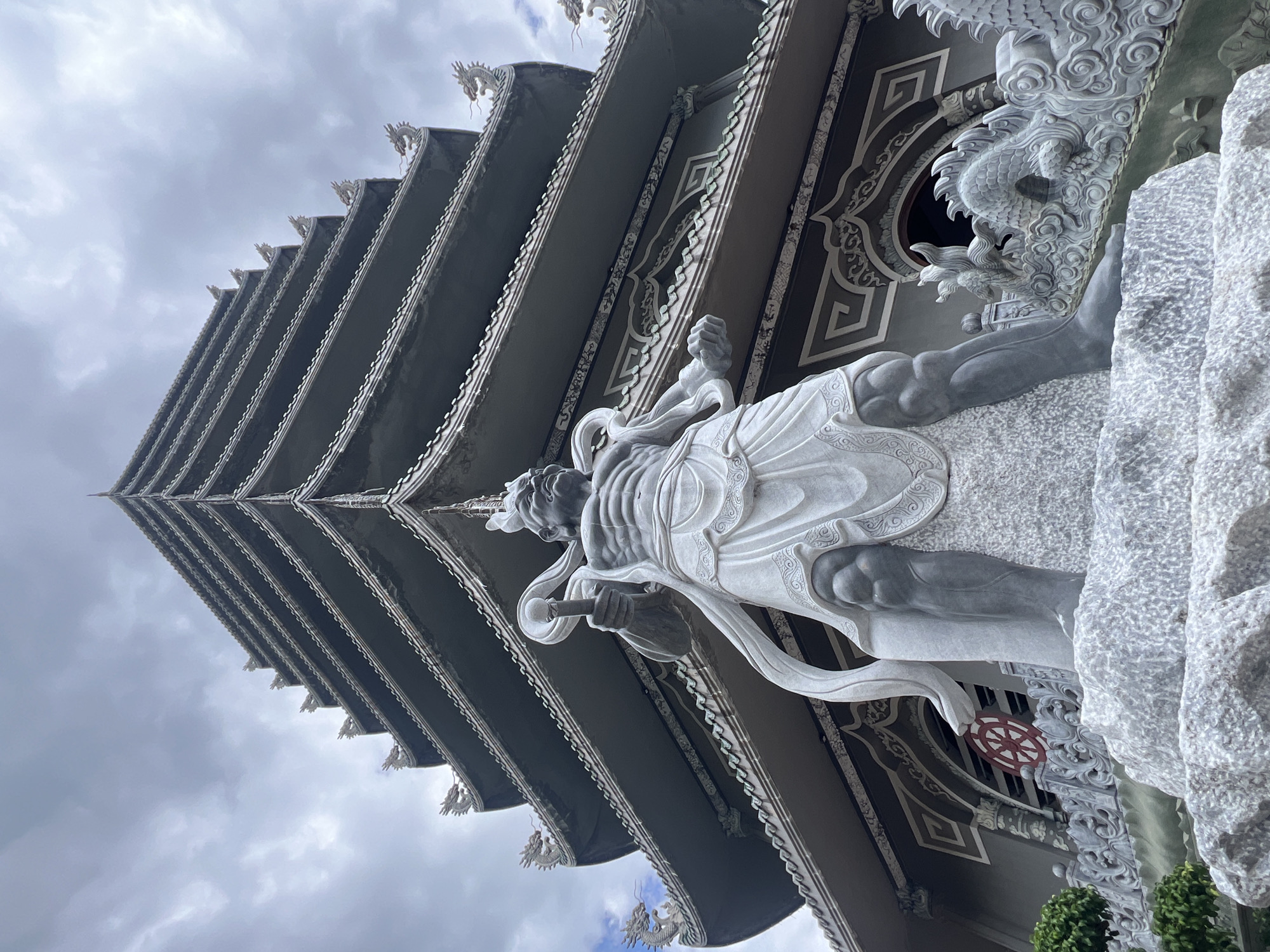 This is a low-angle, full shot of a large, intricately carved statue of a warrior in front of a multi-tiered pagoda in Đà Nẵng, Vietnam. The statue, made of light-colored stone, depicts a muscular figure in a flowing garment, holding a staff in its right hand and a clenched fist in its left. The pagoda behind it is dark gray with ornate eaves, each decorated with small dragon figures. The sky is overcast with clouds. The statue is positioned on a large, rough-hewn stone base, and to its right, there are decorative stone carvings of dragons. A red and white Dharma wheel symbol is visible on the pagoda structure behind the statue. The overall scene suggests a religious or cultural site, likely a temple or pagoda complex, given the architectural style and iconography.