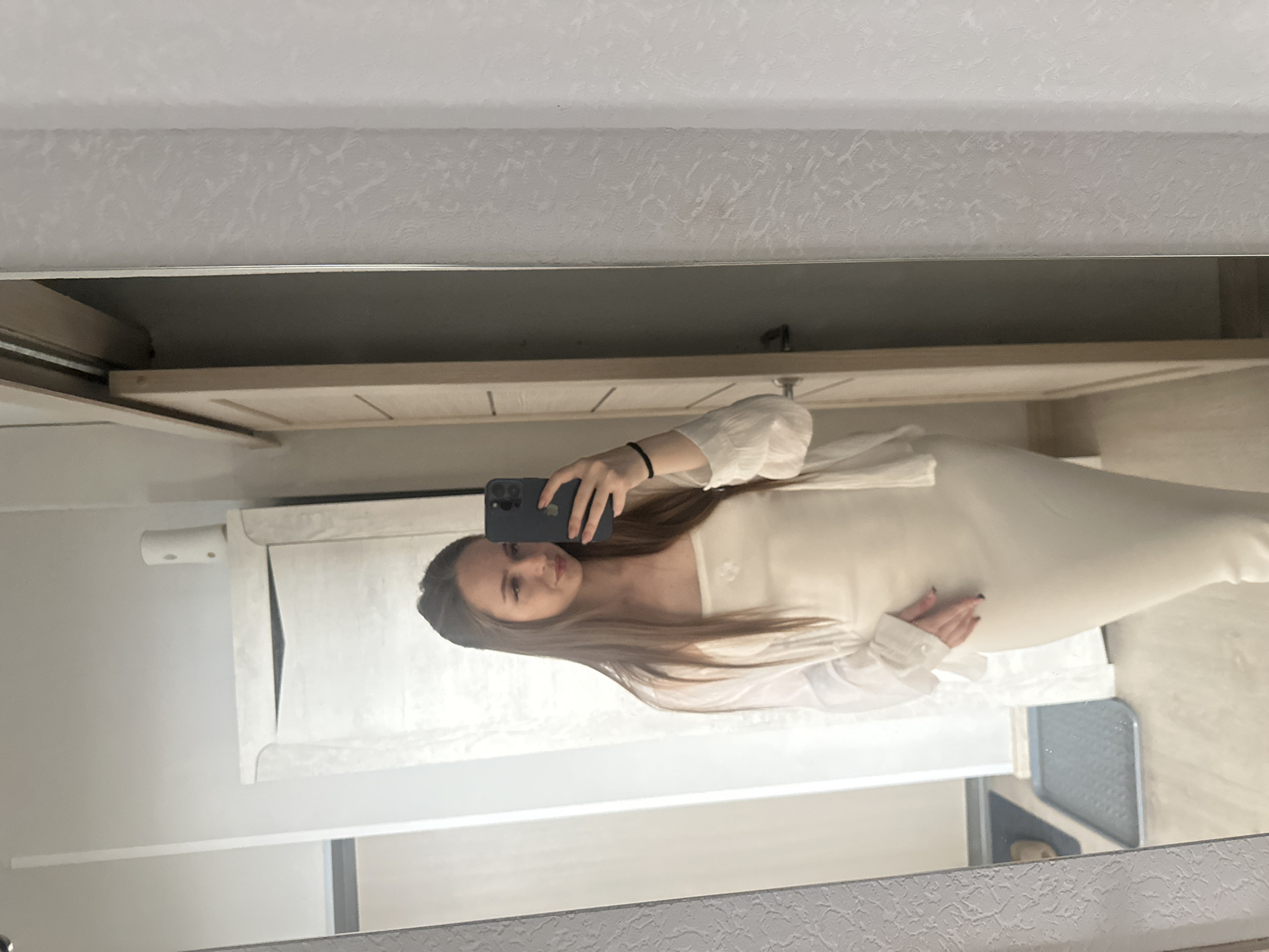 A young woman with long brown hair is standing in front of a mirror, taking a selfie. She is wearing a white strapless dress and a white open-buttoned shirt. Her right hand is on her hip, and her left hand is holding a dark-colored smartphone. The background shows a room with a light-colored door and a patterned wall. The floor appears to be light gray.