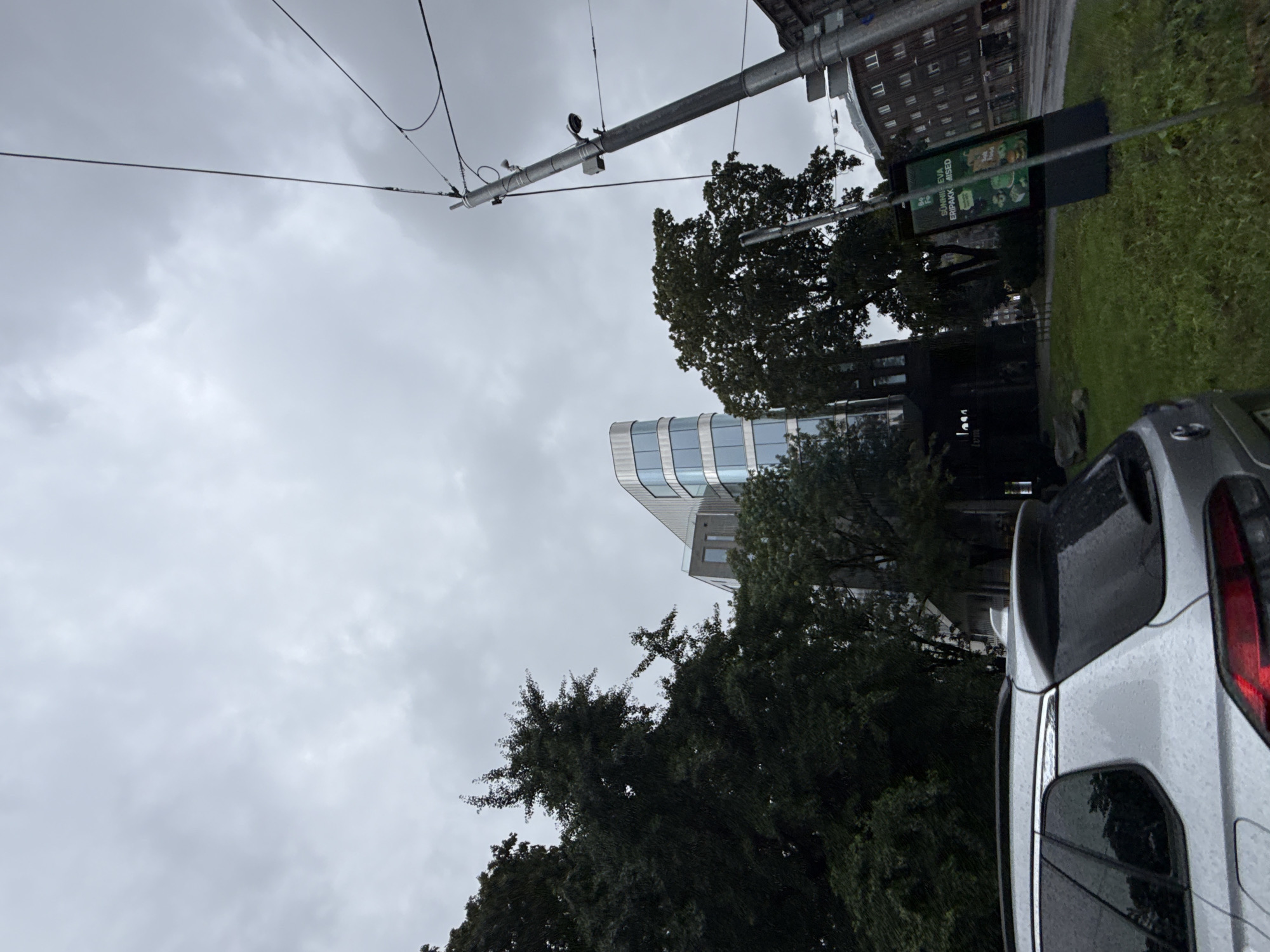 The image captures a street scene in Tallinn, Estonia, under an overcast, possibly rainy sky. In the foreground, the rear of a silver SUV is visible, its surface speckled with water droplets, indicating wet weather. To the left, a dense cluster of dark green trees dominates the frame. Behind the trees, a modern, curved glass-fronted building with a metallic top section rises into the sky. To the right of this building, another older, dark building with multiple windows can be seen. Further to the right, a tall utility pole with electrical wires and a security camera is prominently featured. In the middle ground, there is a digital advertising screen displaying green imagery and text in what appears to be Estonian. The street surface appears wet, and green grass covers the area in front of the advertising screen and between the trees. The overall atmosphere is muted and somber, consistent with inclement weather.