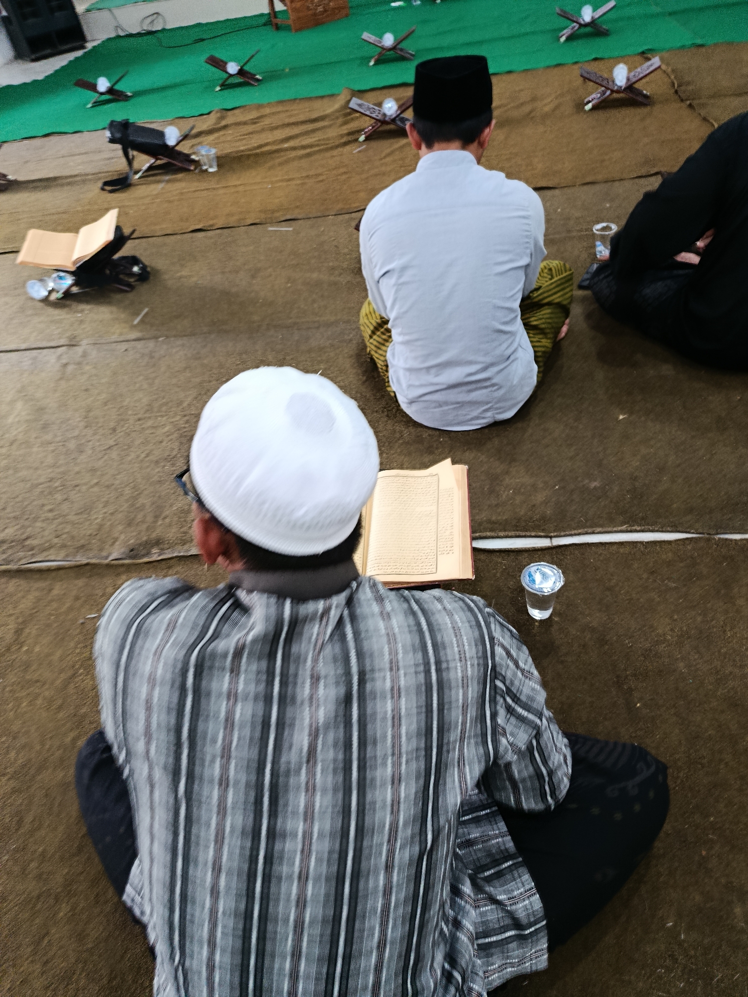 The image depicts an interior scene, possibly a mosque or prayer room, with individuals seated on the floor in a cross-legged position. The floor is covered with a brown carpet, and in the background, a green carpet is visible. Several wooden book stands, each holding an open book, are arranged on both carpets, suggesting a religious gathering or study session.
In the foreground, a person wearing a white kufi and a striped shirt is seated facing away from the camera, with an open book in front of them. To their right, a clear plastic cup containing a clear liquid is placed on the floor. Further in the background, another individual wearing a black kufi and a light-colored shirt is seated, also facing away from the camera. A third person, mostly obscured by shadow and wearing black attire, is seated to the right of the person in the black kufi.
The overall atmosphere appears calm and contemplative. The presence of multiple individuals engaged with religious texts implies an environment dedicated to prayer, study, or worship. The setting is likely indoors, with lighting that creates some shadows, particularly on the right side of the frame. The image does not contain any explicit visual cues to determine its precise location within the city of Bondowoso, Indonesia, beyond the possibility of it being a place of worship or religious education.