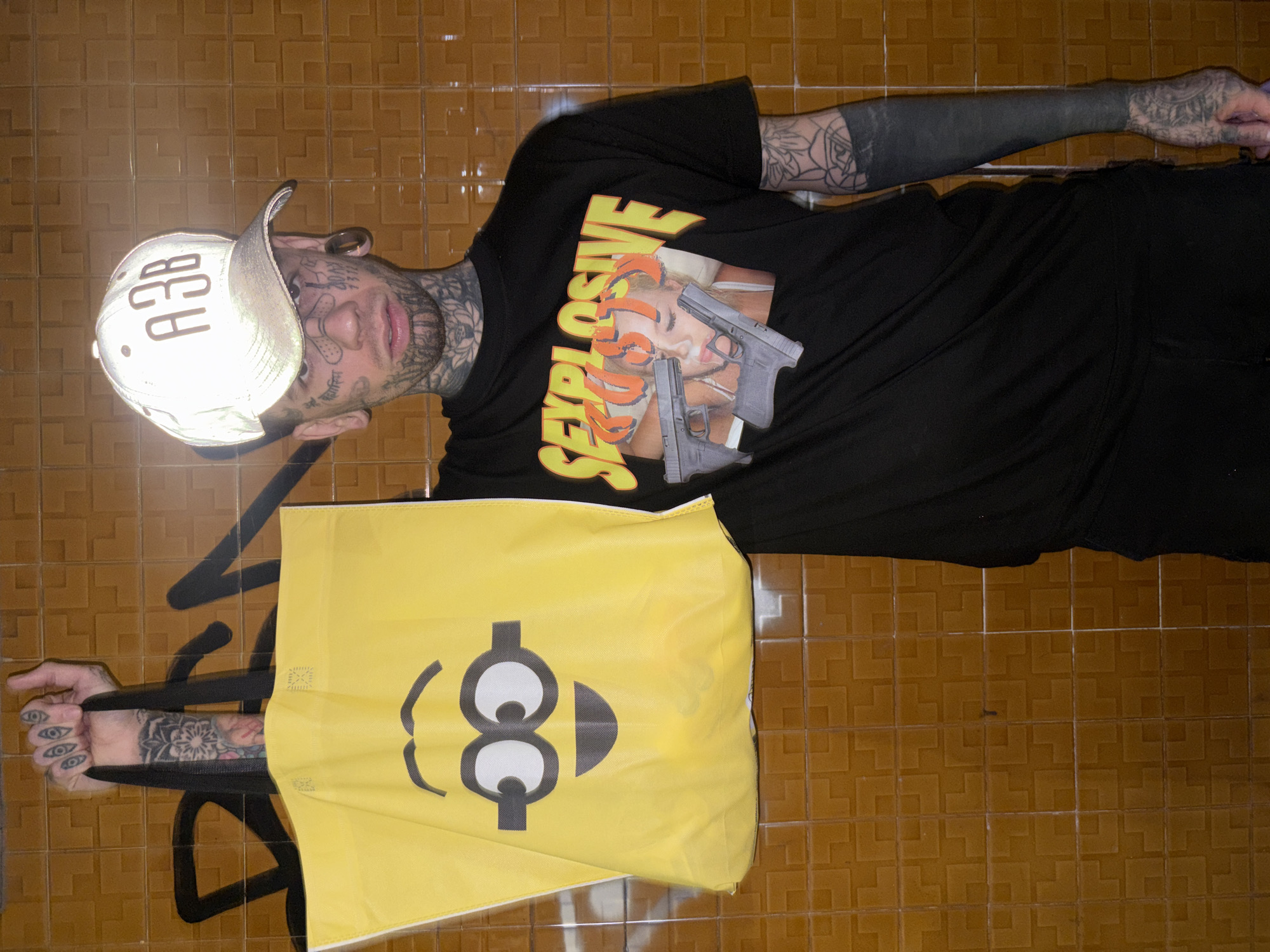 A person is lying on a tile floor. The person wears a baseball cap with "A3B" written on it, a black t-shirt with "SEXPLOSIVE" printed on it along with an image of a woman and two handguns, and black pants. The person is heavily tattooed, including on the face and arms. A yellow shopping bag with a cartoon face printed on it is draped over the person's lower body. The tile floor is brown with a geometric pattern. The location is not clear from the image.
