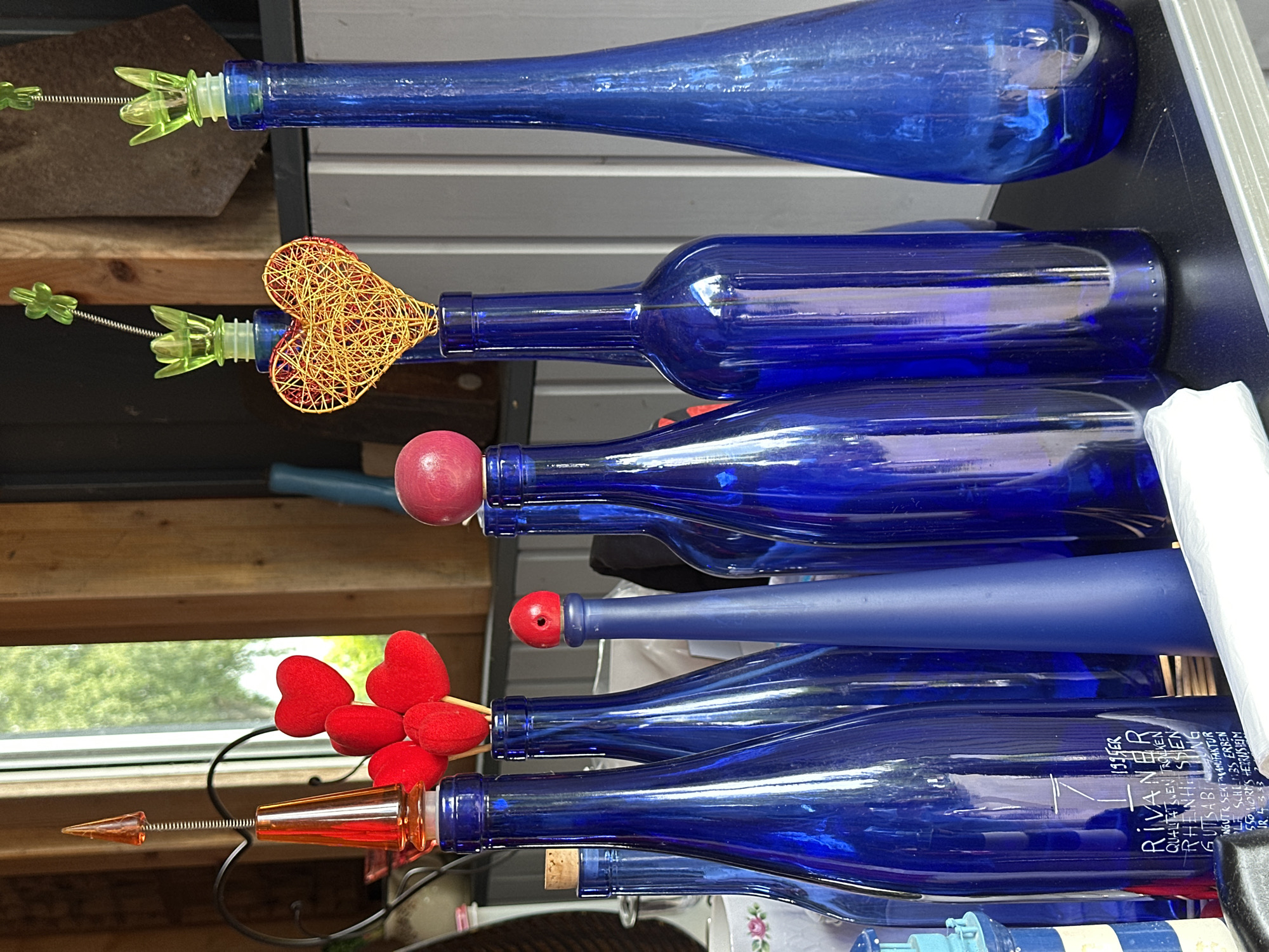The image depicts an indoor scene in Deinste, Germany, showcasing approximately ten cobalt blue glass bottles of various shapes and sizes, arranged on a black, reflective surface. Several bottles are fitted with decorative stoppers or inserts: a translucent orange, pointed, helical stopper; four red fabric hearts on individual sticks; a small red wooden spherical stopper; a yellow wire-wrapped heart; and two translucent green plastic stoppers shaped like stylized flowers. A natural cork stopper is also visible in one bottle. One bottle displays a white printed label with text identifying it as "1995ER RIVANER QUALITÄTSWEIN TROCKEN RHEINHESSEN GUTSABFÜLLUNG." An opaque, matte blue, conical object is positioned among the other bottles. The background features a window revealing green foliage, vertical wooden planks, a dark metal heart-shaped stand, and white vertical wall paneling. A white ceramic bowl with a rose pattern and a white plastic item are present in the lower-left background and foreground, respectively. No human figures are depicted.