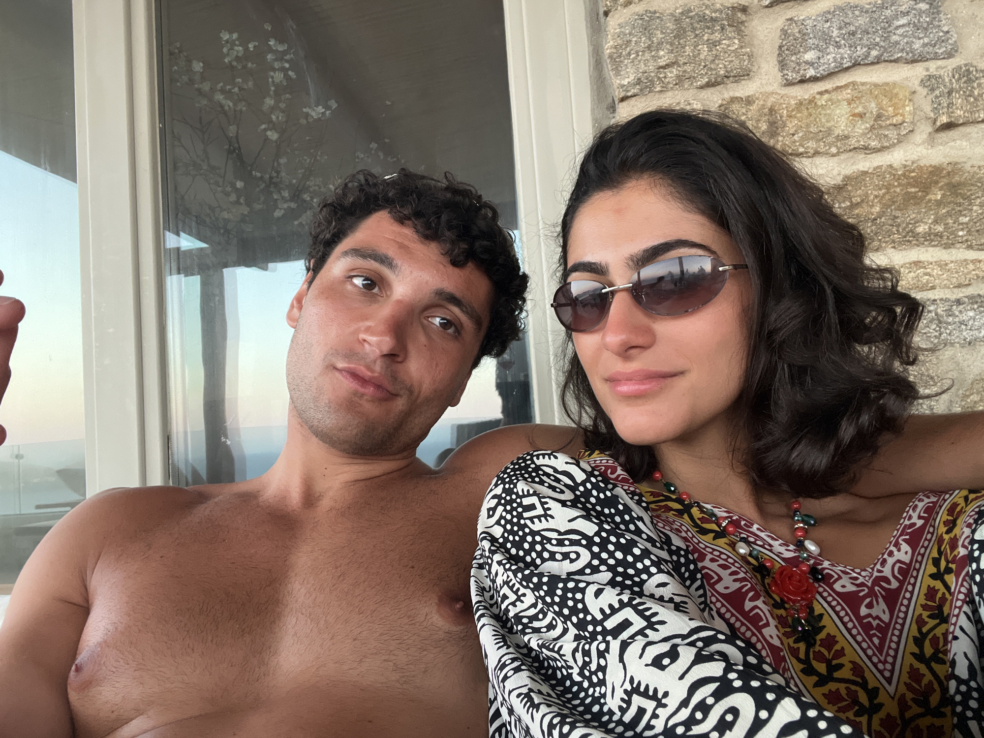A shirtless man with dark, curly hair and a woman wearing sunglasses and a colorful patterned dress are posing for a selfie outdoors. They are seated, with the man on the left and the woman on the right. The background shows a stone wall and a window with a reflection of a tree. The scene suggests a relaxed, possibly vacation setting, likely in a coastal area of Greece given the architecture and lighting.