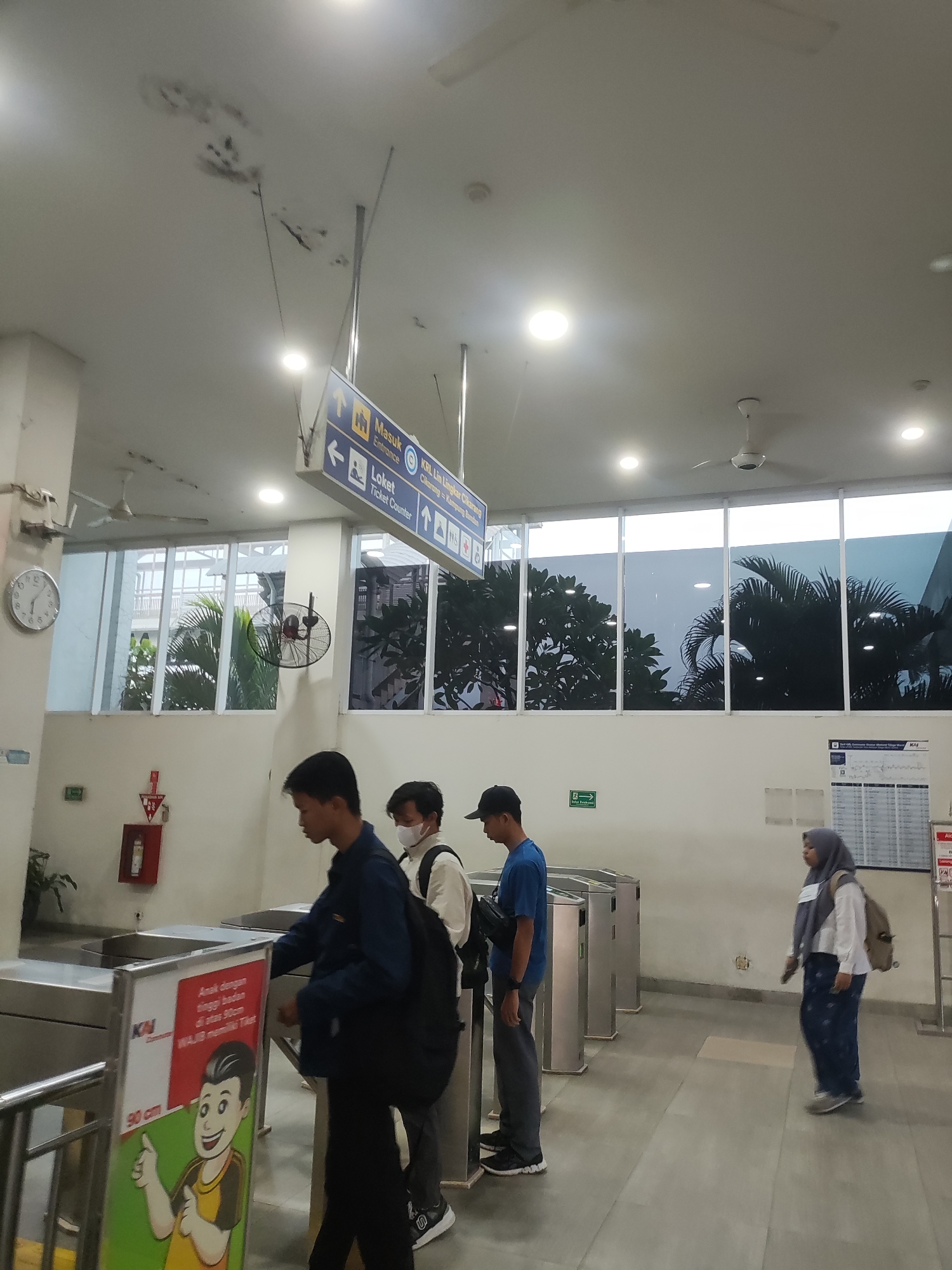 The image depicts an indoor public transportation station, likely a train or KRL (Commuter Line) station, with elements suggesting it is located in Telagamurni, Indonesia, based on signage.
In the foreground, three individuals are visible. On the left, a young man wearing a dark blue jacket and black pants is passing through a turnstile, looking down. A backpack is slung over his right shoulder. Next to him, a person wearing a white shirt, black vest, and white face mask, also with a backpack, is standing. To the right, another young man in a blue t-shirt and grey pants is walking away from the turnstiles, with a black bag across his chest. Further to the right, a woman wearing a grey hijab, white shirt, and patterned pants is walking towards the turnstiles.
The environment features a white ceiling with lights and ceiling fans, and a large window looking out onto trees and foliage, suggesting a tropical climate. Multiple turnstiles for entry and exit are present. Signage in Indonesian is visible, including directional signs pointing towards "Masuk" (Entrance) and "Loket" (Ticket Counter), and indicating "KRL Lin Lingkar Cikarang-Kampung Bandan". A safety sign with an emergency exit symbol is also visible on the wall. A clock with a white face and black hands is mounted on the left wall. A fire extinguisher is mounted on the wall near the clock. A large poster featuring a cartoon character and text is placed near the turnstiles. The floor is tiled with a light grey pattern.
