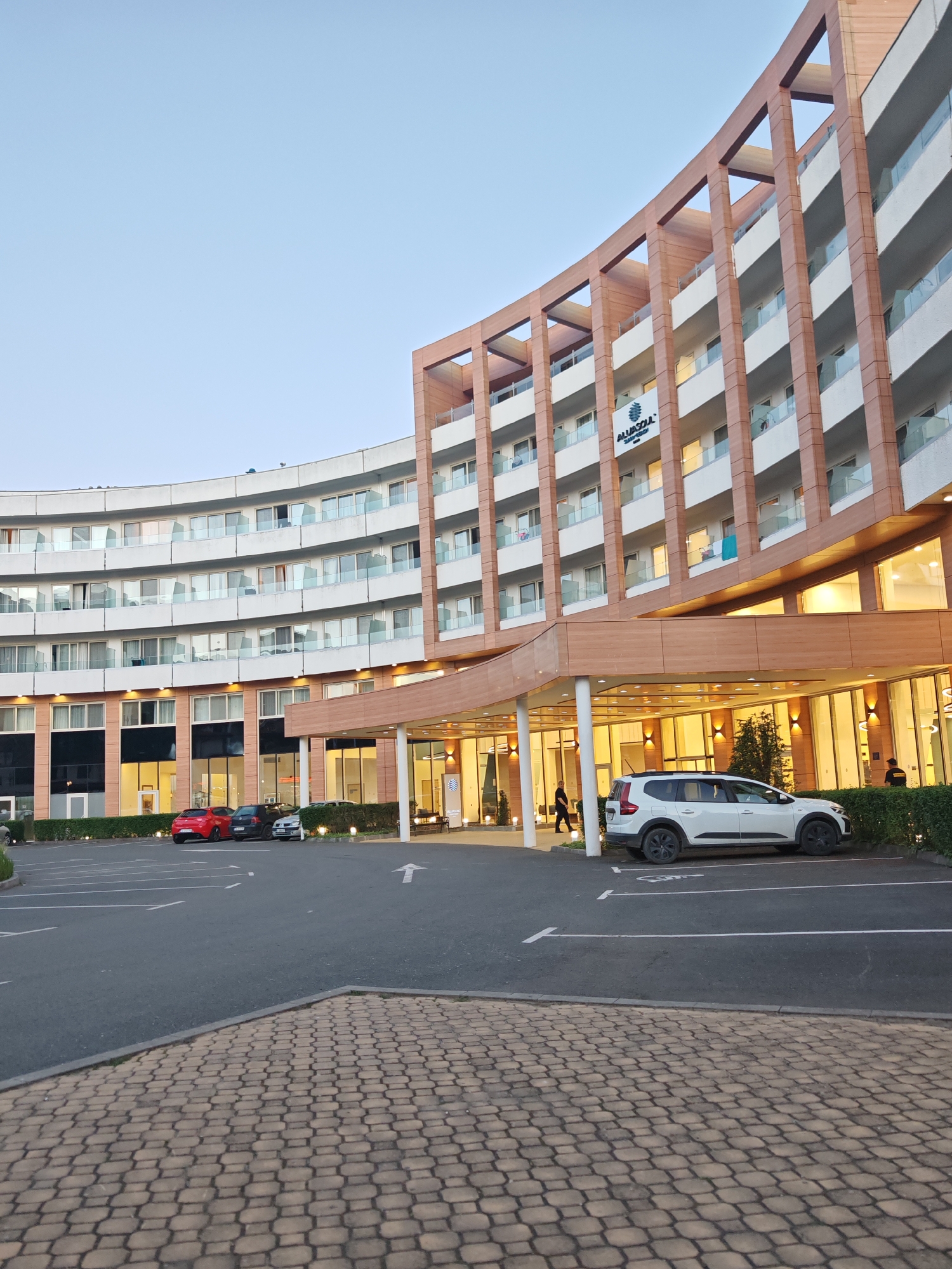 The image shows the All Inclusive Hotel Allasoul Sunny Beach in the city of Nesebar, Bulgaria. The hotel is a curved, multi-story building with white balconies and a wooden exterior. A white SUV is parked under the covered entrance. In the parking lot in front of the hotel, a red car, a dark gray car, and a white car are visible. An arrow is painted on the pavement, indicating the direction of traffic. A woman walks towards the entrance of the hotel. The sky is pale blue. Paved bricks are visible in the foreground.
