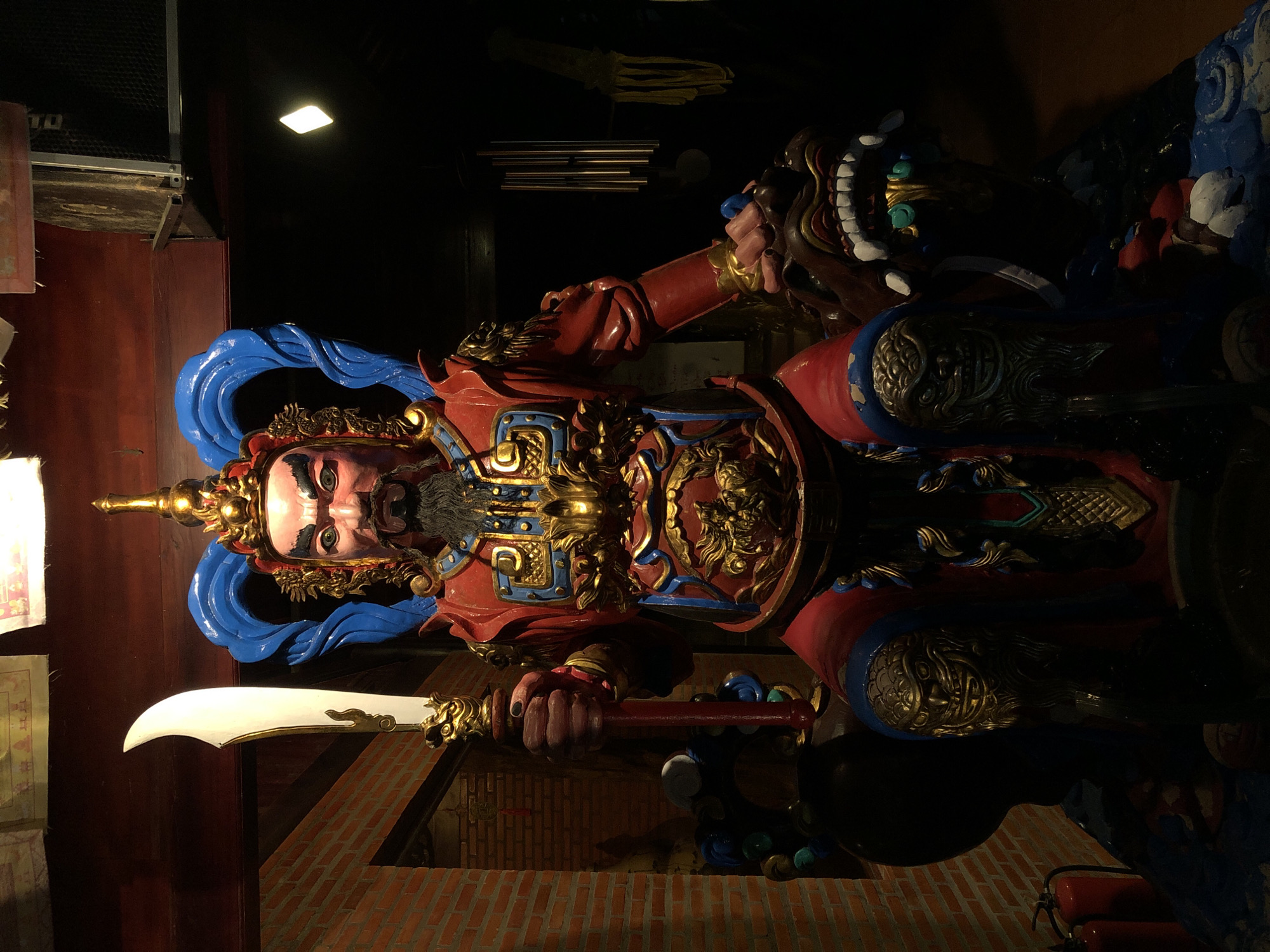 This is a photograph of a large, ornate statue of a warrior, likely a deity or historical figure from East Asian culture. The statue is brightly colored with reds, blues, and gold, and is adorned with intricate details. The warrior has a stern expression, a long black beard, and is wearing elaborate armor and a headdress. He is holding a large, curved sword in his right hand. To the warrior's right, there is a grotesque, dragon-like head with sharp teeth, also ornately decorated. The statue is positioned in what appears to be an indoor setting, with a dark background and a brick-like surface visible at the bottom. Above the statue, there are some dark objects, possibly part of the ceiling or decorative elements. The lighting suggests it is nighttime or the area is dimly lit. The overall impression is one of power and grandeur. The location context provided is Mũi Né, Vietnam, which might suggest the statue is part of a temple or cultural attraction in that area. There is no visible text in the image.
