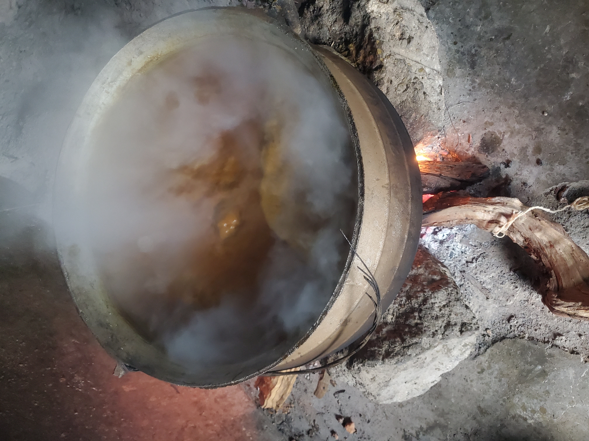 A large, dark pot with a silver rim sits over an open fire, with plumes of white steam rising from its contents. The pot is supported by rocks and appears to be cooking food over burning wood. The scene suggests an outdoor cooking area, possibly in a rural or less developed part of Jos, Nigeria. The ground is rough and unpaved, with ash and debris from the fire visible. The lighting is dim, suggesting it might be nighttime or early morning. No people are visible in the image.
