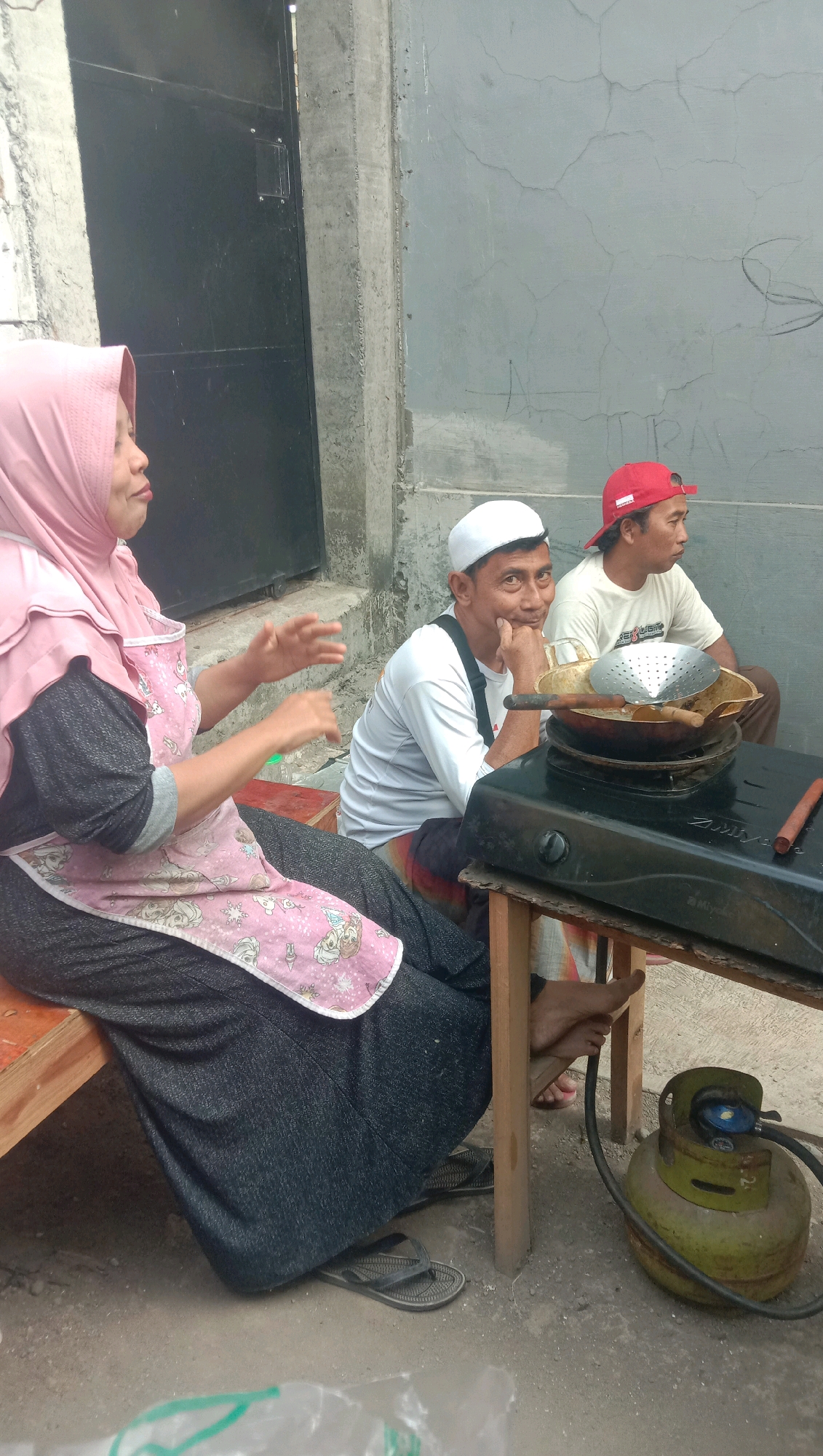 In Karang Anyar, Indonesia, three individuals are captured in an outdoor scene, possibly near a residential or commercial establishment, with a weathered concrete wall in the background and a black metal door to the left. A woman wearing a pink hijab and a pink apron adorned with cartoon characters is seated on a wooden bench, gesturing with her hands. To her right, two men are also seated. One man, wearing a white cap and a white long-sleeved shirt, rests his chin on his hand, looking towards the camera. Beside him, another man in a red cap and a light-colored t-shirt is looking to the right. A portable gas stove with a pan and cooking utensils is positioned on a makeshift wooden table in front of the men, with a green gas cylinder connected to it. The ground is covered with dirt and gravel, and a plastic bag is visible in the foreground.