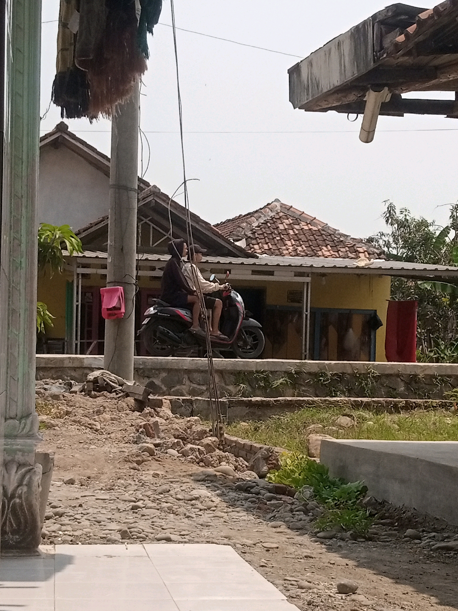 The image depicts a street scene in what appears to be a rural or suburban area of Boyoteluk, Indonesia. In the foreground, a rough, rocky path leads towards a concrete structure. A utility pole stands prominently on the left side of the path, with some laundry hanging from it.
Behind the pole, two individuals are riding a black motorcycle. The person in the rear is wearing a dark hijab and a long-sleeved top, while the person in the front wears a light-colored long-sleeved top and a baseball cap. The motorcycle is parked beside the utility pole.
Further in the background, a yellow house with a red-tiled roof is visible. The house has a covered porch area and some windows. Trees and foliage surround the house, suggesting a lush environment. The overall impression is of a typical day in a quiet Indonesian community.
