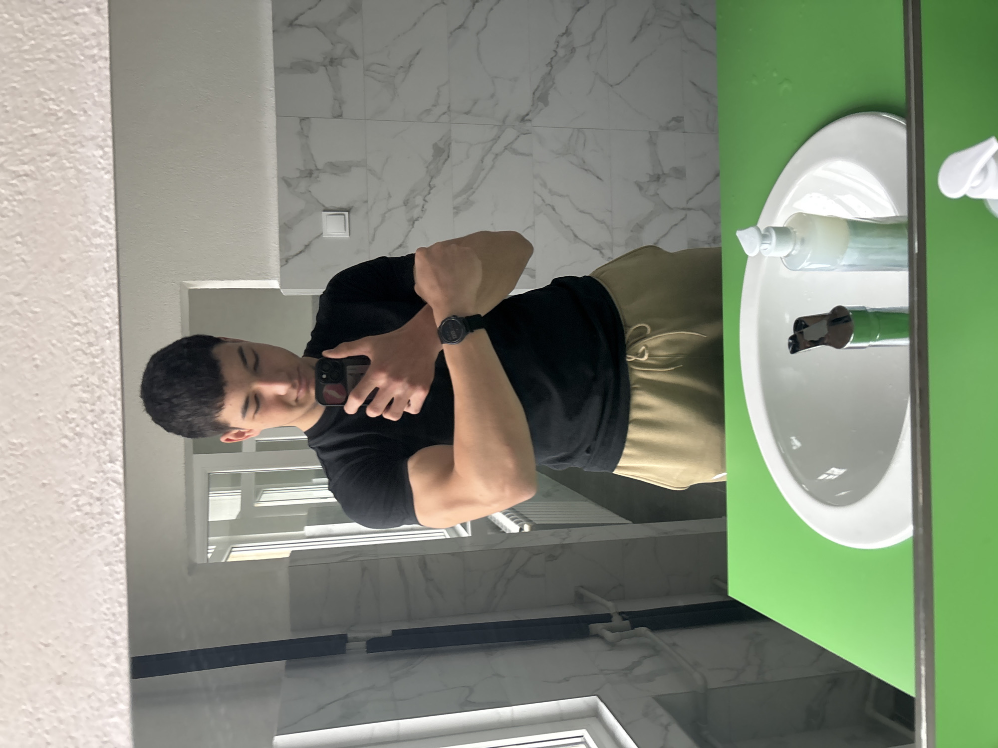 A young man stands in a bathroom, reflected in a mirror as he takes a selfie with his smartphone. He is wearing a black t-shirt, beige pants, and a black watch. The bathroom features white marble-patterned wall tiles. A white sink is visible, with a green surround, reflecting cleaning products and a tap. Windows are visible in the reflection, and the location is likely Zlatograd, Bulgaria, inferred from the context of other images.