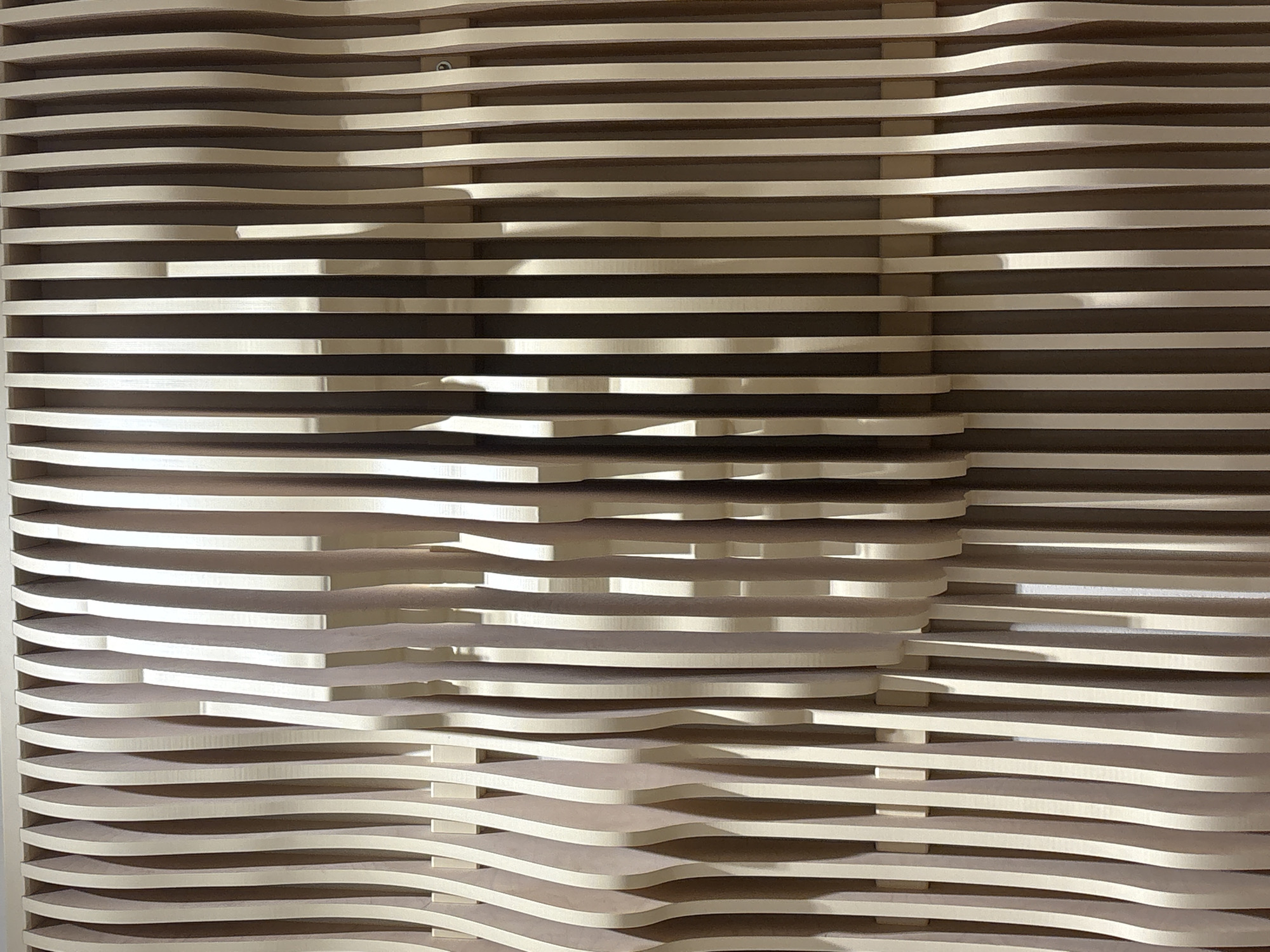 The image shows a close-up, eye-level view of a textured wall made of vertical wooden slats. The slats are of varying widths and depths, creating a wave-like pattern with shadows and highlights. The wood appears to be a light to medium brown color, with some slats having a darker, more muted tone. The overall effect is one of depth and organic form. The background behind the slats is indistinct, appearing to be a darker, possibly recessed area. The image is well-lit, emphasizing the texture and form of the wooden structure. There are no people or any other discernible objects in the frame. The location is described as Unknown, Unknown.