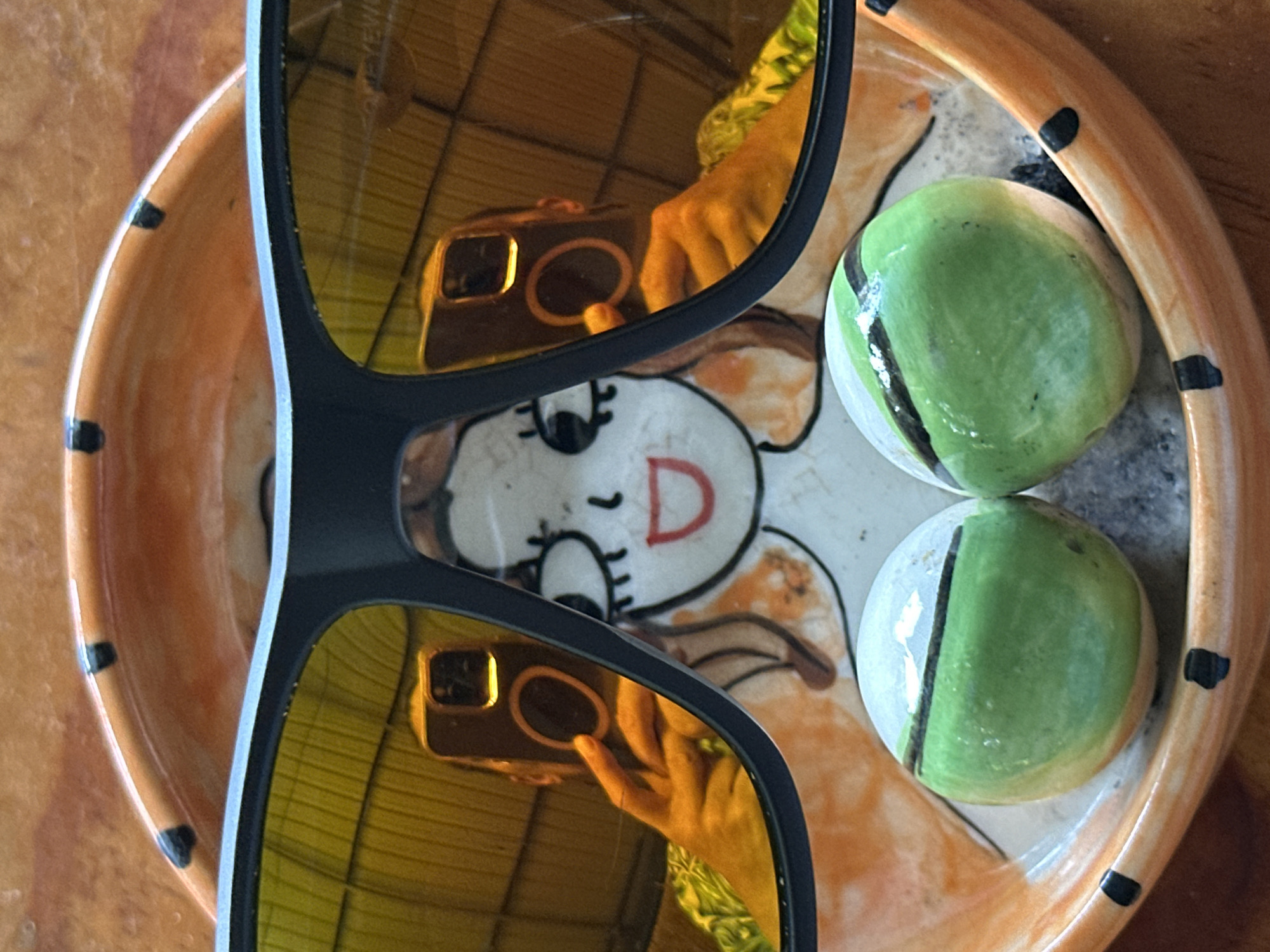 The image shows a still life composition on a wooden surface. An orange-rimmed dish with black dot accents contains two green and white decorative orbs. The dish features a central design of a cartoon-like figure with the letter "D" prominently displayed. A pair of black-framed sunglasses with amber-colored lenses are positioned on the dish, reflecting the image of a person holding a smartphone. The location of the image is ตำบลเกาะพะงัน, Thailand.