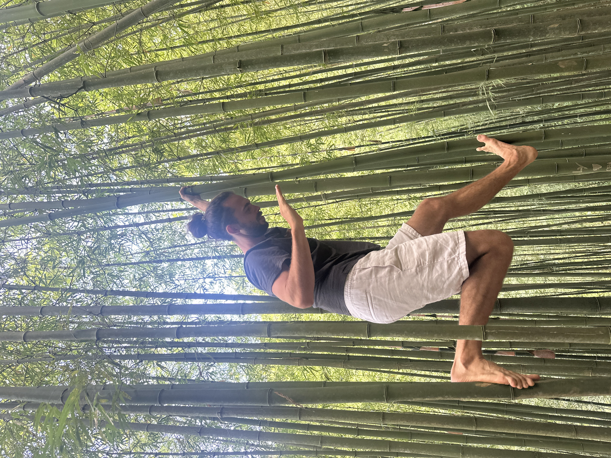 A man in a dark t-shirt and light-colored shorts is lying horizontally in a bamboo forest. His right leg is bent at the knee and his foot is touching the bamboo stalks above him. His left leg is straight and his foot is touching the bamboo stalks below him. His right arm is raised up towards his head. The bamboo forest is dense and the bamboo stalks are green. This photograph was taken in the city of Ngã Ba Kim, Vietnam.