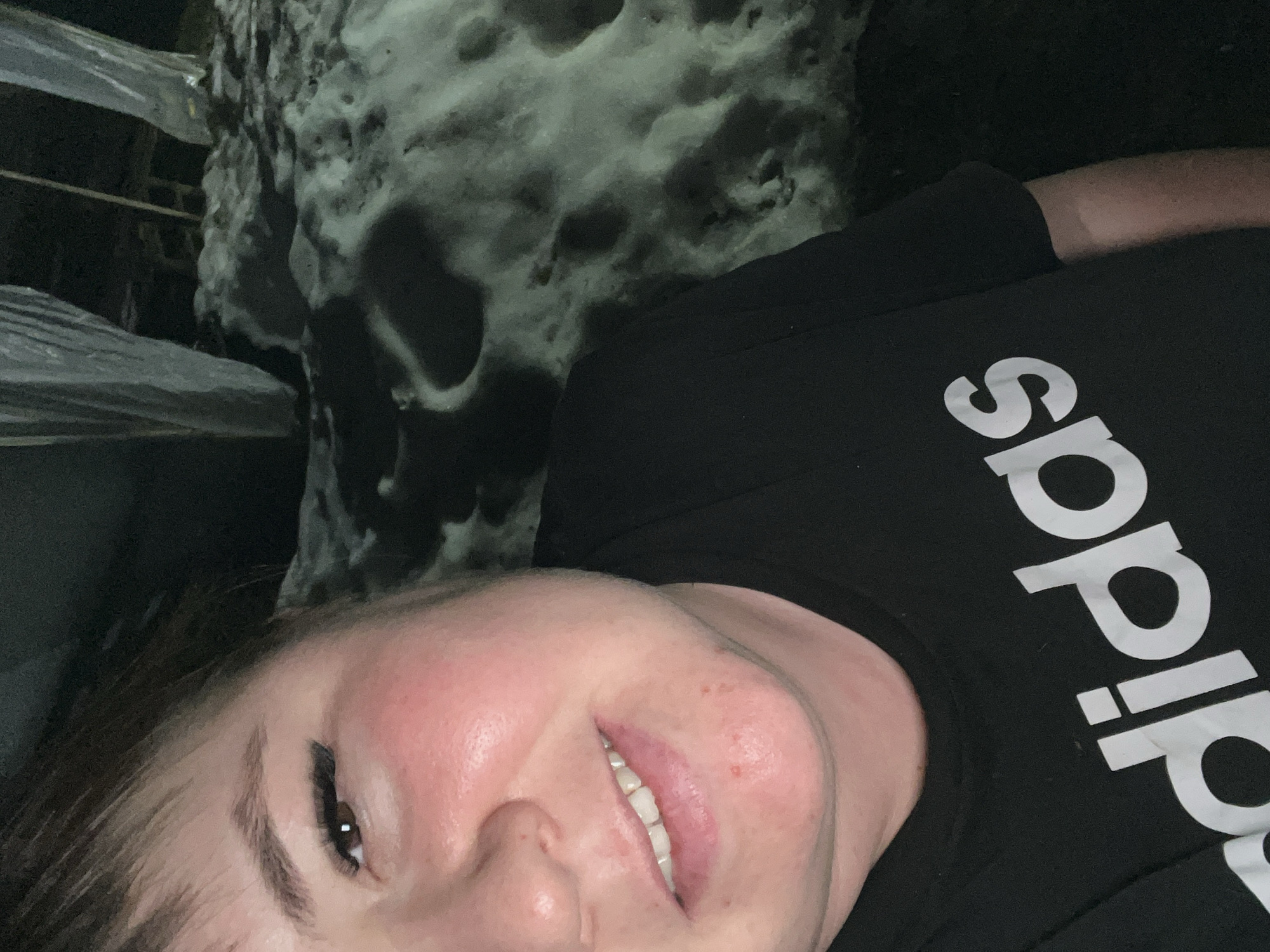 A person wearing a black Adidas t-shirt is looking at the camera and smiling, appearing slightly out of breath. The person is positioned in front of a rock structure, possibly a rock formation, which might indicate they are outdoors in a natural setting or in a constructed rock garden-like area. Based on the prompt, the location may be Chaikovsky, Russia, though the image doesn't inherently show this. There is some kind of fence or railing next to the rock.