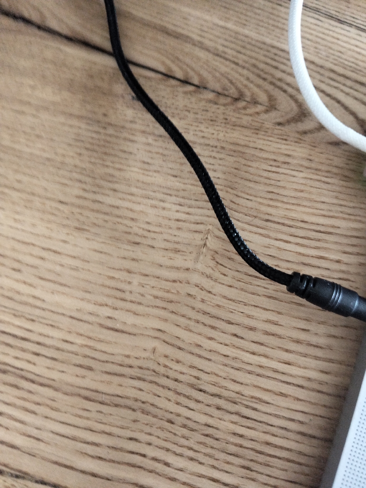 A close-up, overhead shot shows a black cord with a black connector plugged into a device, resting on a wooden surface. To the right of the black cord, a white cord is visible, also on the wooden surface. The wooden surface is light brown with a visible grain pattern. The scene appears to be indoors, on a desk or table. There are no people visible.