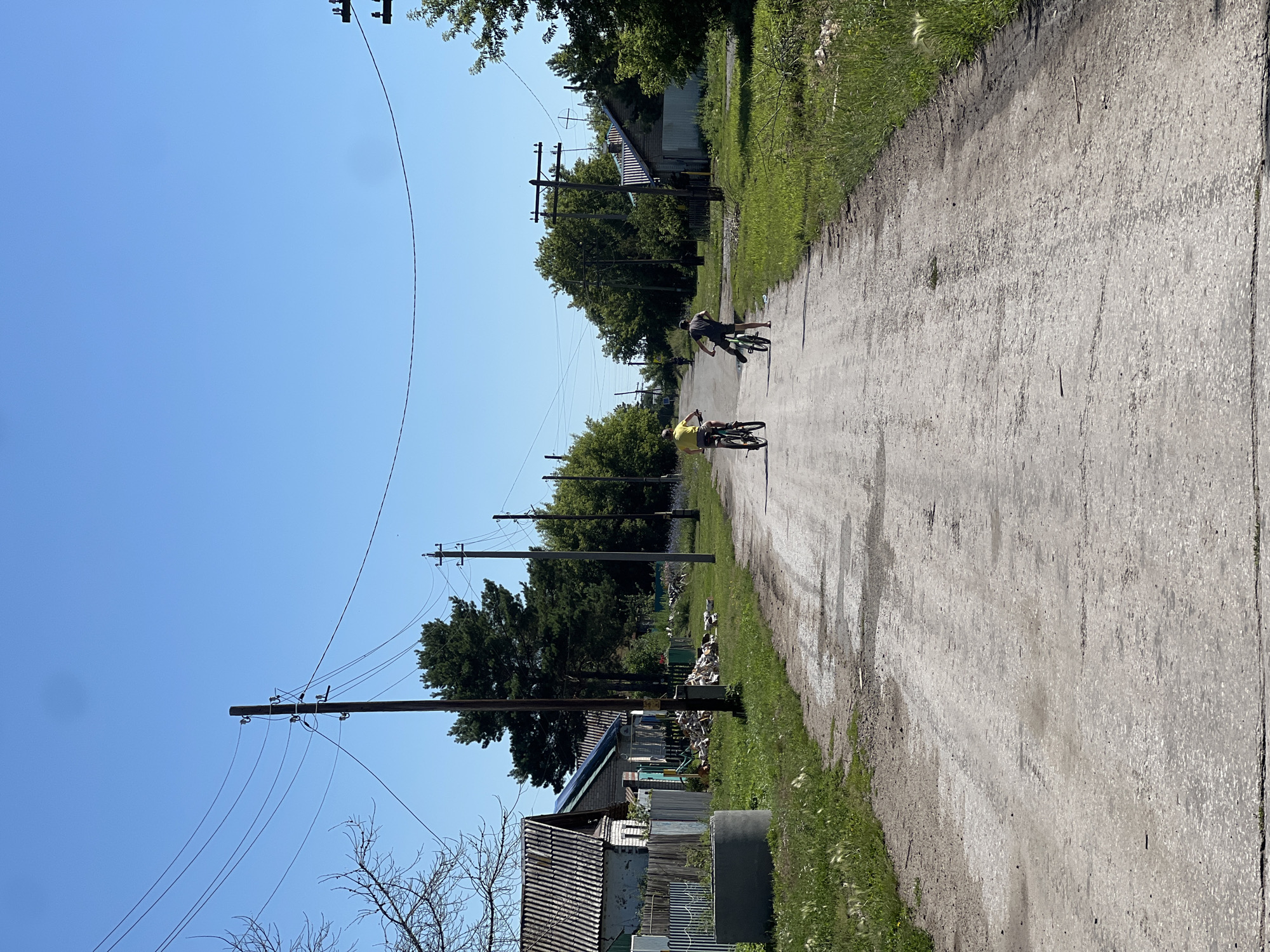 In Pivkino, Russia, on a dirt road lined with houses and trees, two children ride bicycles under a clear blue sky. The road is dusty and uneven, with tire tracks visible. On the left side of the road, there are several houses with corrugated roofs, a wooden fence, and a concrete well. On the right side, there are more houses and trees. Power lines stretch across the sky, supported by wooden poles. The scene appears peaceful and ordinary, depicting a typical summer day in a rural Russian village.