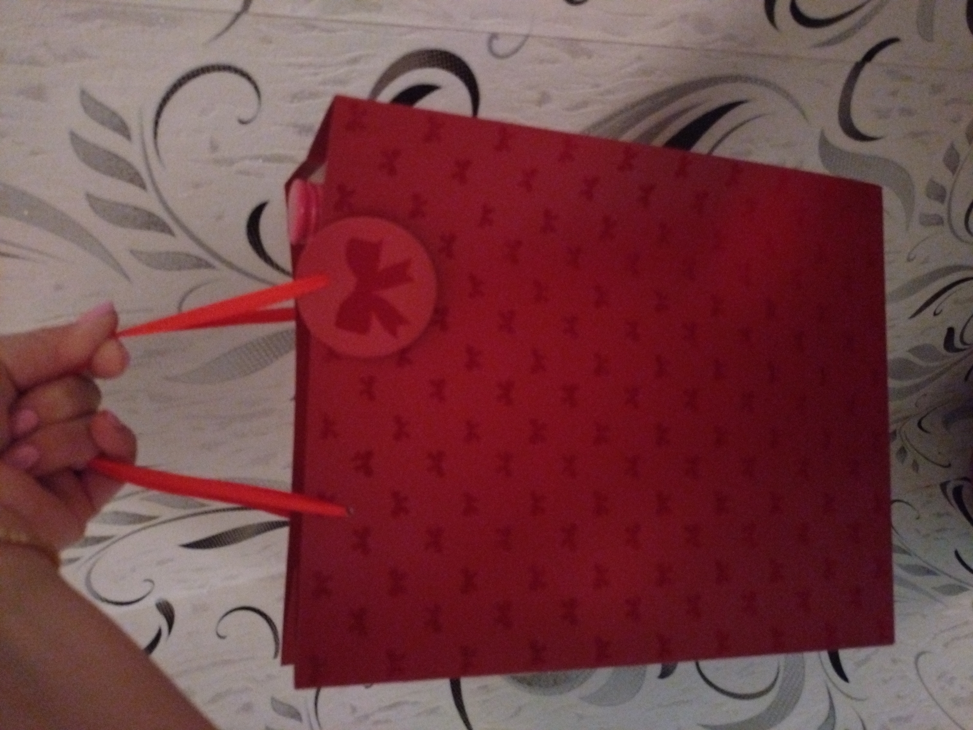 A fair-skinned hand with light pink manicured fingernails is observed from the wrist up, grasping the red ribbon handles of a vertical rectangular gift bag. The bag is a solid red color, patterned with numerous subtle, darker red bow shapes uniformly distributed across its surface. A circular red tag, embossed or printed with a larger central bow design, hangs from the right handle. Inside the bag, a small, pink, cylindrical object is partially visible near the top right edge. The background consists of a wall covered in wallpaper featuring an elaborate swirling and curvilinear pattern in black, various shades of grey, and white on a light, textured surface. No information within the image indicates the city of Stara Zagora, Bulgaria, or any specific geographic location.