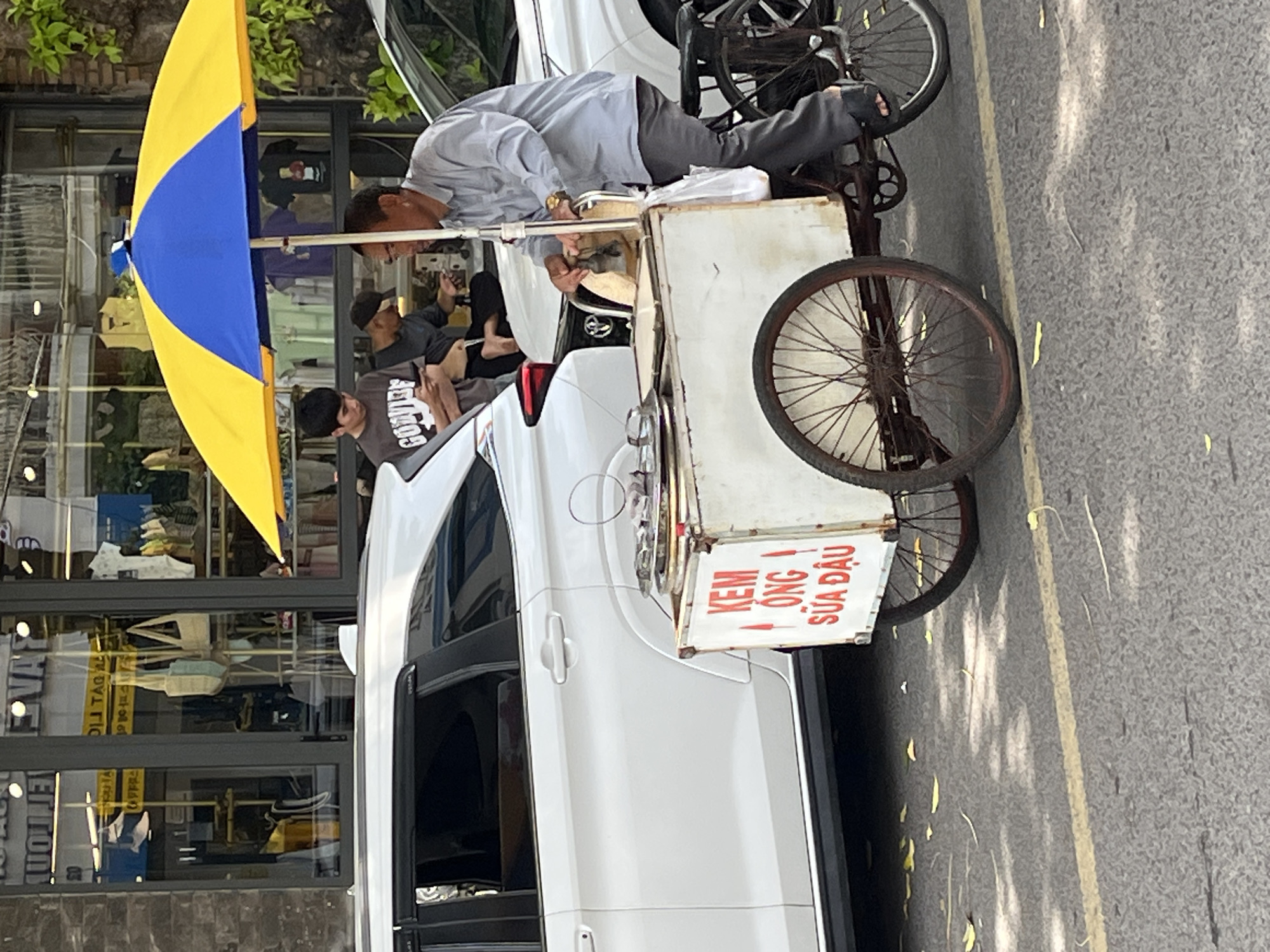 An outdoor daytime scene captures a street vendor on a bicycle cart selling ice cream in Nha Trang Ward, Vietnam. The vendor, an adult male wearing a light blue long-sleeved shirt and dark pants, sits on his cart and appears to be interacting with the ice cream equipment. Above the cart, a blue and yellow umbrella provides shade. Two younger individuals are visible behind the cart. One, a male wearing a gray t-shirt, is seated and looking at a mobile phone. Another male, wearing a dark cap, is standing and looking down. The white ice cream cart has red lettering that reads "KEM ONG SUA DAU." The cart is positioned next to a white car. In the background, a storefront with large glass windows displays clothing items. The street is paved, and fallen yellow leaves are scattered on the ground. The lighting suggests it is a sunny day with dappled sunlight on the street.
