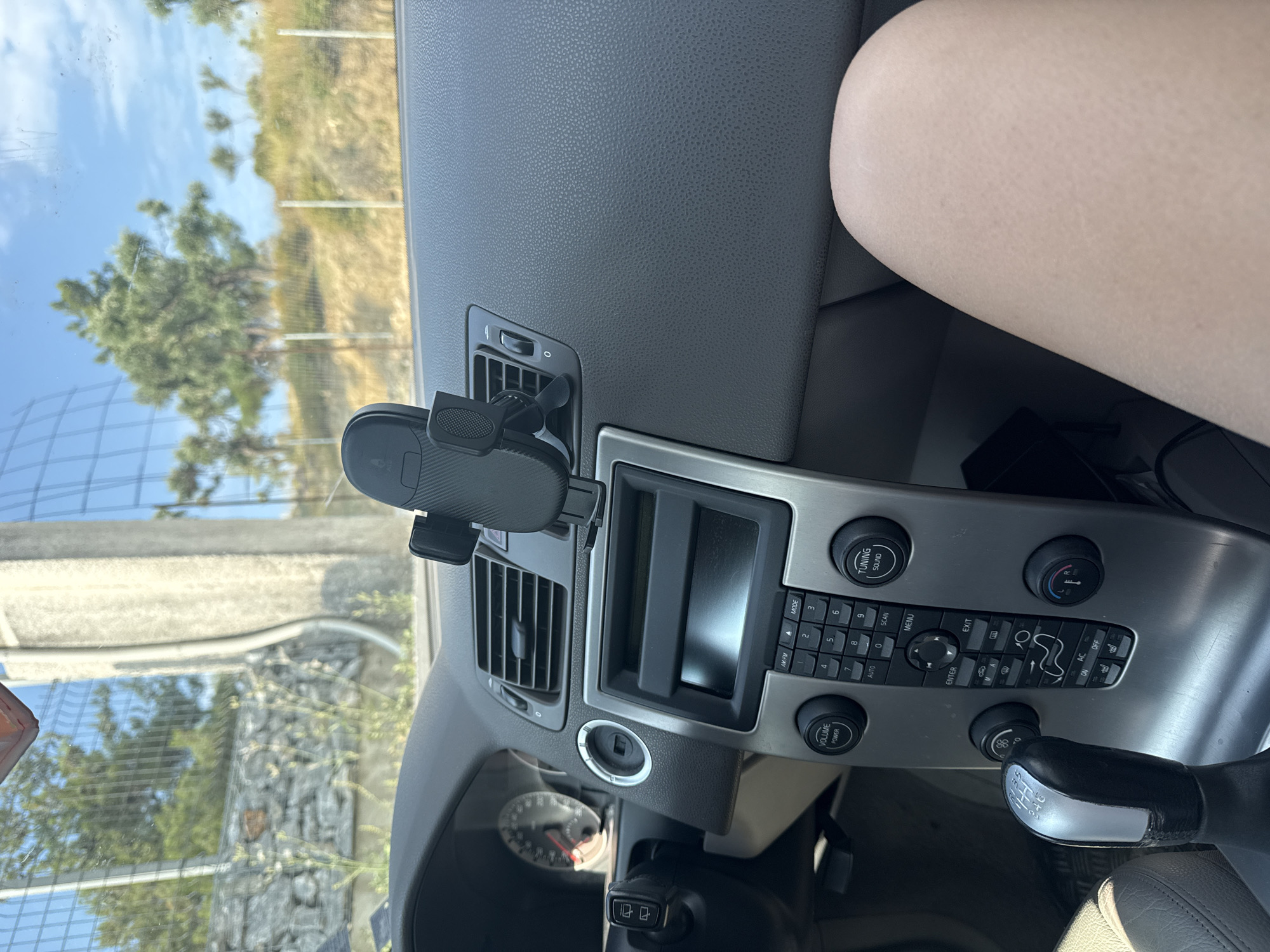 The image is a medium shot of the interior of a car, focusing on the dashboard and a person's leg. The dashboard features a car mount holding a phone, air vents, a car stereo, and various buttons and controls including a gear shift. Outside the car, a fence and trees are visible against a bright sky, suggesting an outdoor, possibly rural, environment. The scene is static, with no discernible action or interaction occurring. The lighting is bright and natural, indicating daytime.