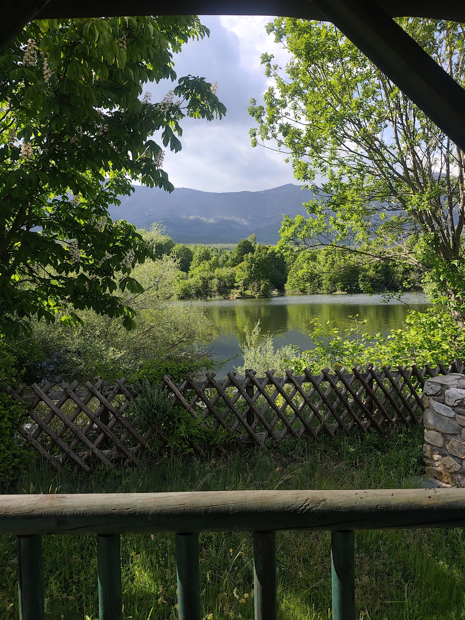 The image shows a view of a lake and mountain range framed by trees and a wooden structure. The lake appears calm and reflects the green trees lining its banks. A lattice fence runs along the foreground. In the distance, a mountain range is visible under a cloudy sky. The scene is lush and green. Location details: Unknown, Unknown.