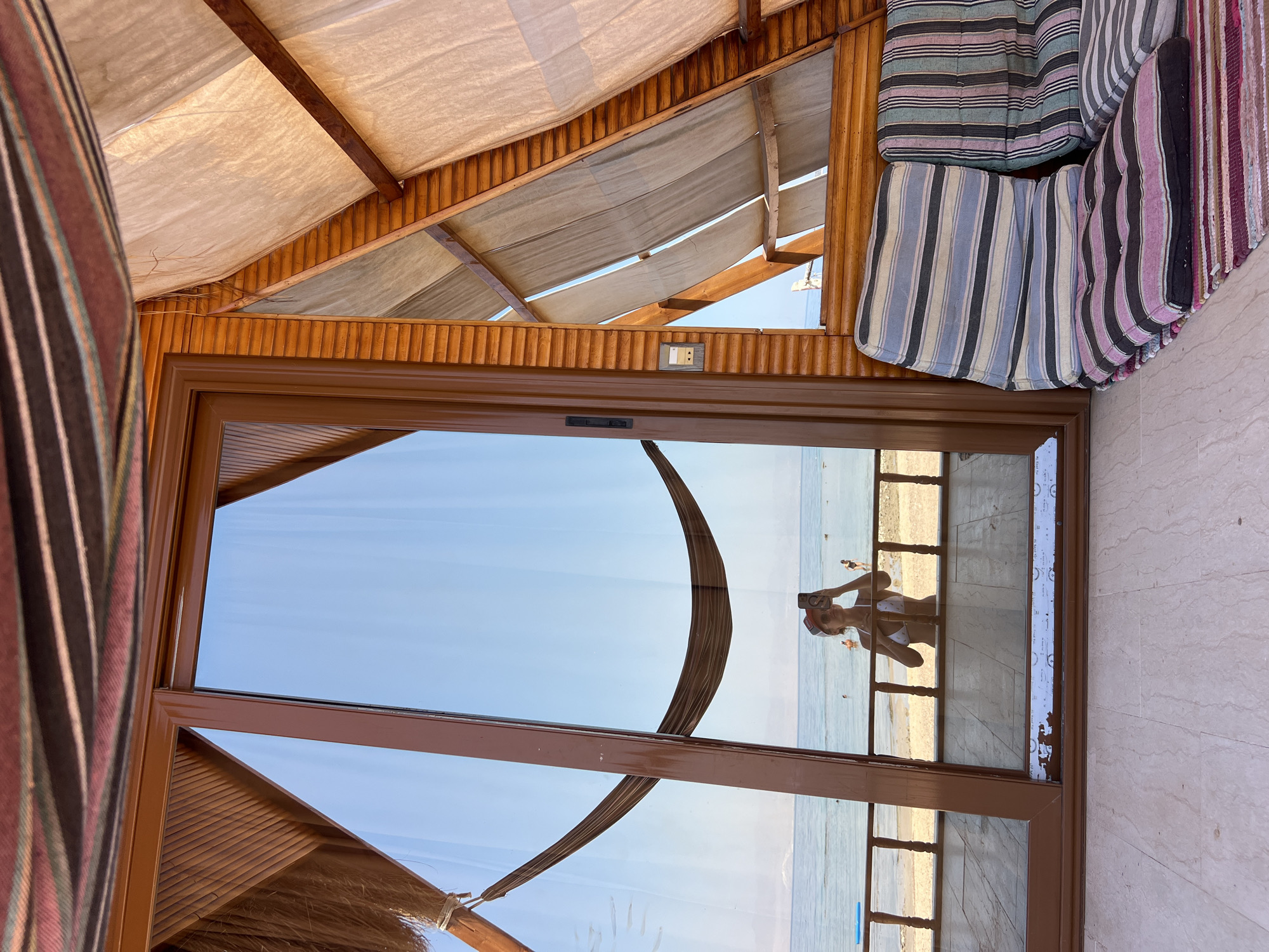 The image depicts a beachside scene in Unknown, Egypt, viewed from the interior of a triangular wooden structure. In the foreground, on the right, are two striped floor cushions and a striped rug. A large glass door dominates the left side of the frame, reflecting the exterior. In the reflection, a person wearing a bikini and a cap is visible holding a phone and taking a selfie. Beyond the person and the railing in the reflection, a beach and the calm sea are visible under a clear blue sky. Further in the background, several people are in the water. The interior of the structure shows wooden walls and beams, with white fabric draped as a canopy.