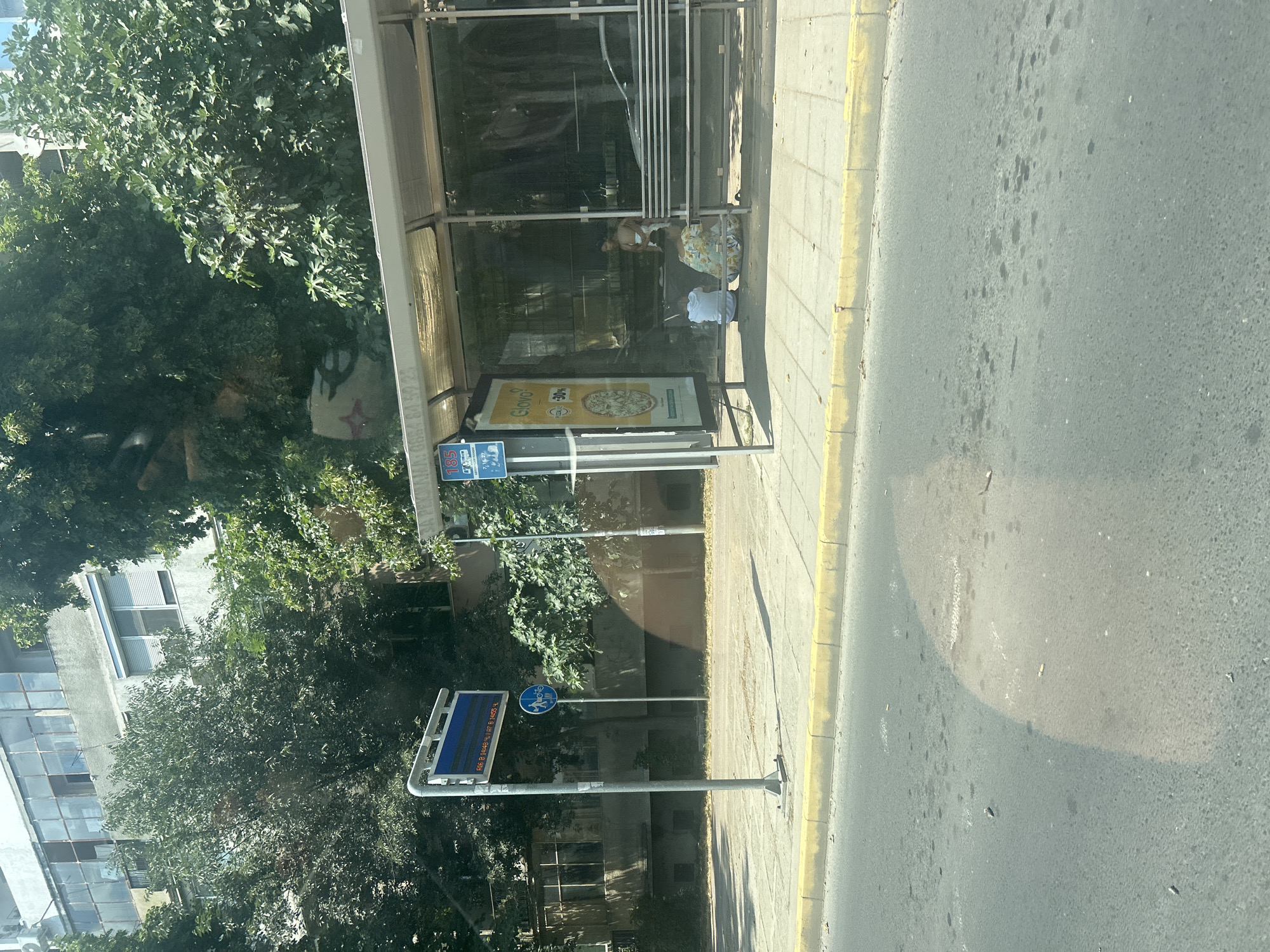 The image depicts a street scene in Plovdiv, Bulgaria, likely near a bus stop. In the foreground, the asphalt road shows patches of dark stains and scattered cigarette butts, indicating urban wear and tear. A yellow curb separates the road from a concrete sidewalk.
On the sidewalk, a bus stop shelter with a transparent roof and glass panels stands. Inside the shelter, a metal bench is visible. Three people are present at the bus stop. One individual, appearing to be a child, stands near the back of the shelter. Two adults are seated on the sidewalk facing away from the camera, one wearing a floral-patterned dress and the other in a white shirt.
To the left of the bus stop, a tall metal pole supports a blue digital display sign showing what appear to be bus route information or arrival times, and below it, a circular blue sign with white symbols of a bicycle and a pedestrian. Behind the bus stop and sidewalk, a dense line of trees and a multi-story building with balconies and windows form the backdrop. The overall lighting suggests it is daytime.