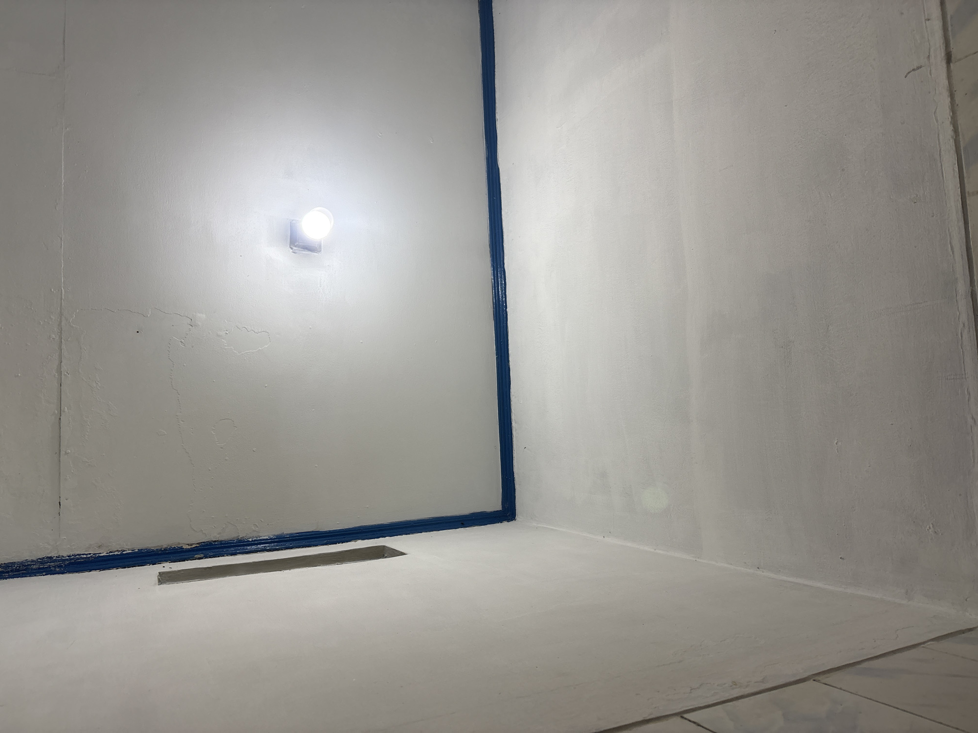 A dimly lit interior corner of a room is captured from a low angle. The walls and ceiling are painted white, showing some signs of wear and minor cracks. A dark blue painted line runs along the junction of the walls and ceiling. In the upper right section of the ceiling, a single, bright light bulb is illuminated in a square fixture, casting a strong glow that washes out some of the ceiling detail. A vertical gray element is visible on the left wall. The image provides no information about Palimanan, Indonesia, or any specific context beyond the visual elements presented.