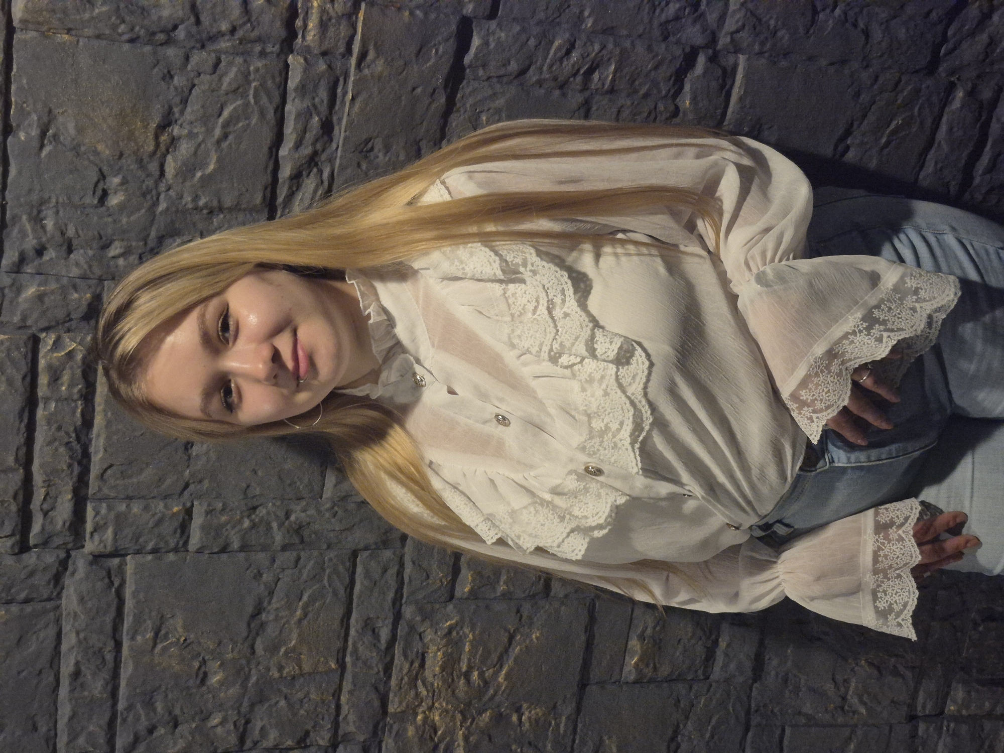 A young woman with blonde hair, wearing a white ruffled blouse and blue jeans, stands against a dark stone wall. She has hoop earrings and a lip piercing. The lighting suggests an indoor setting, possibly a themed restaurant or a nightclub. There is no identifiable landmark or context that places her specifically in Chaikovsky, Russia.