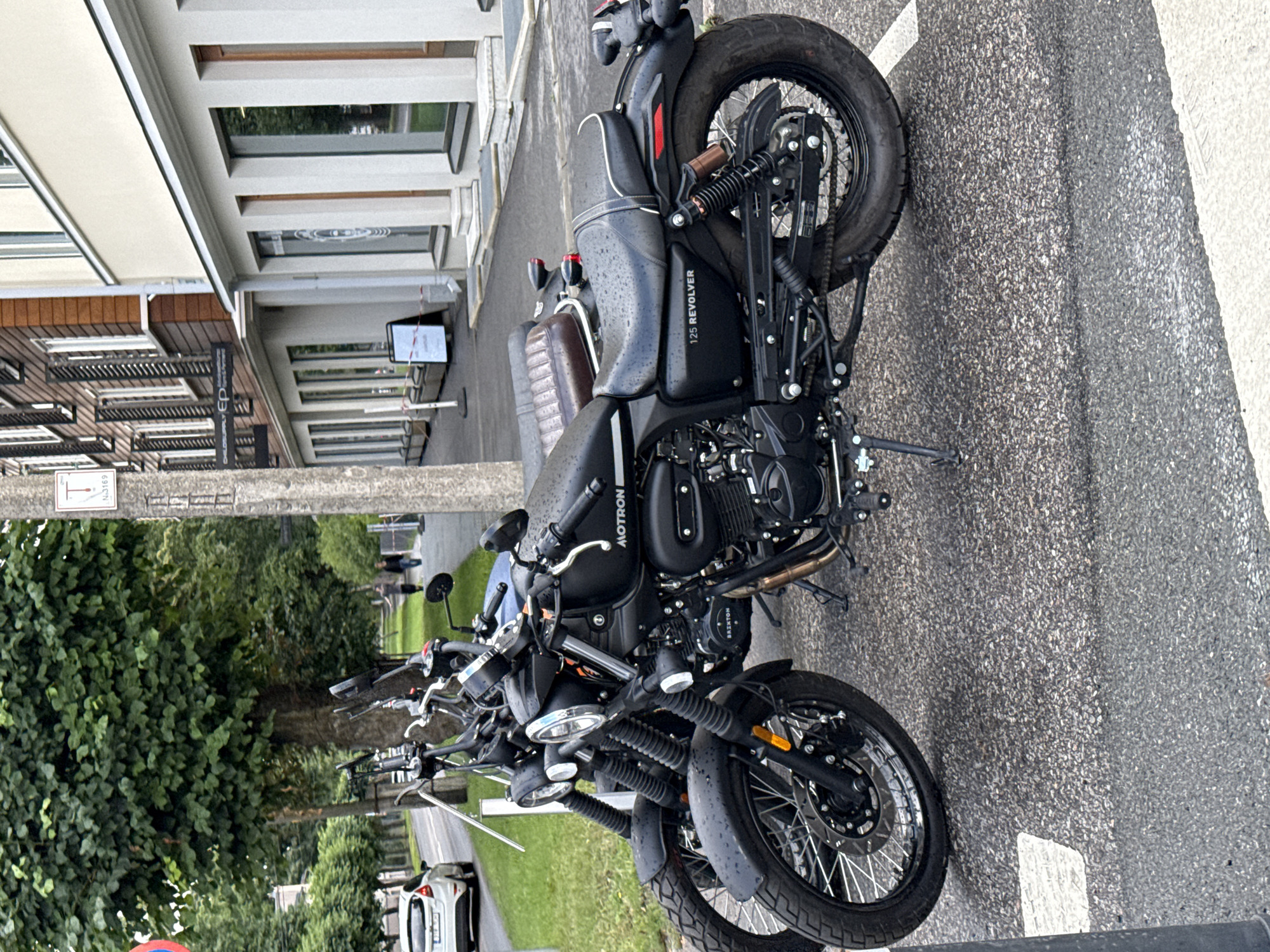 In Tallinn, Estonia, on a street lined with trees and buildings, two black motorcycles are parked. The motorcycle in the foreground has "MOTRON" and "125 REVOLVER" visible on its body. Both motorcycles are wet, suggesting recent rain. A white car is seen driving on the road in the background. The scene is set against a backdrop of urban architecture, with a concrete pole and signs also visible.