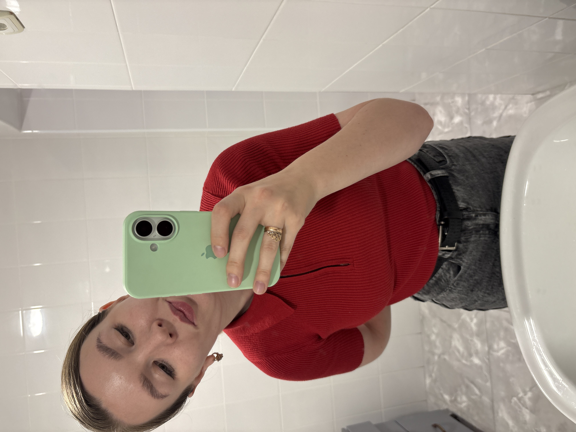 A woman is taking a selfie in a bathroom. She is wearing a red top, gray jeans with a black belt, and a ring. She's holding a light green iPhone. The bathroom has white tiled walls and a white sink. No information is available regarding the location of the bathroom within the city of Izhevsk, Russia.