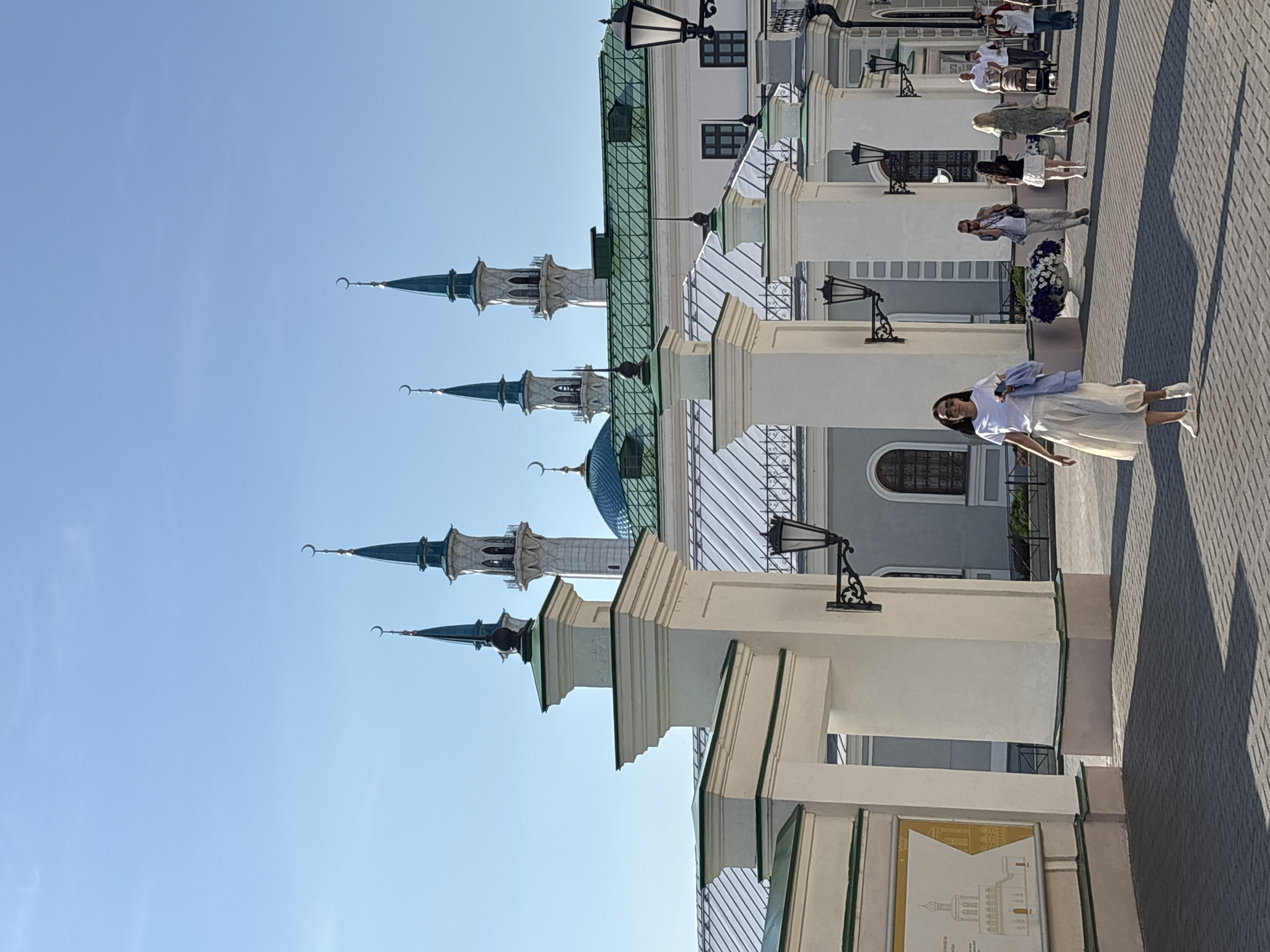 In the city of Kazan, Russia, a bright sunny day illuminates the Kul Sharif Mosque complex. The camera captures a view of the mosque's distinctive turquoise domes and slender minarets, adorned with crescent moons. In the foreground, a white gate structure with decorative pillars stands. Several people are walking in the courtyard. A woman in a white t-shirt and a flowing cream-colored skirt is walking towards the camera, holding a blue shirt in her left hand. Other individuals, including couples and a person with a baby stroller, are visible in the background, walking away from the camera. The ground is paved with gray bricks, and the shadows of the pillars and people are cast on the ground.