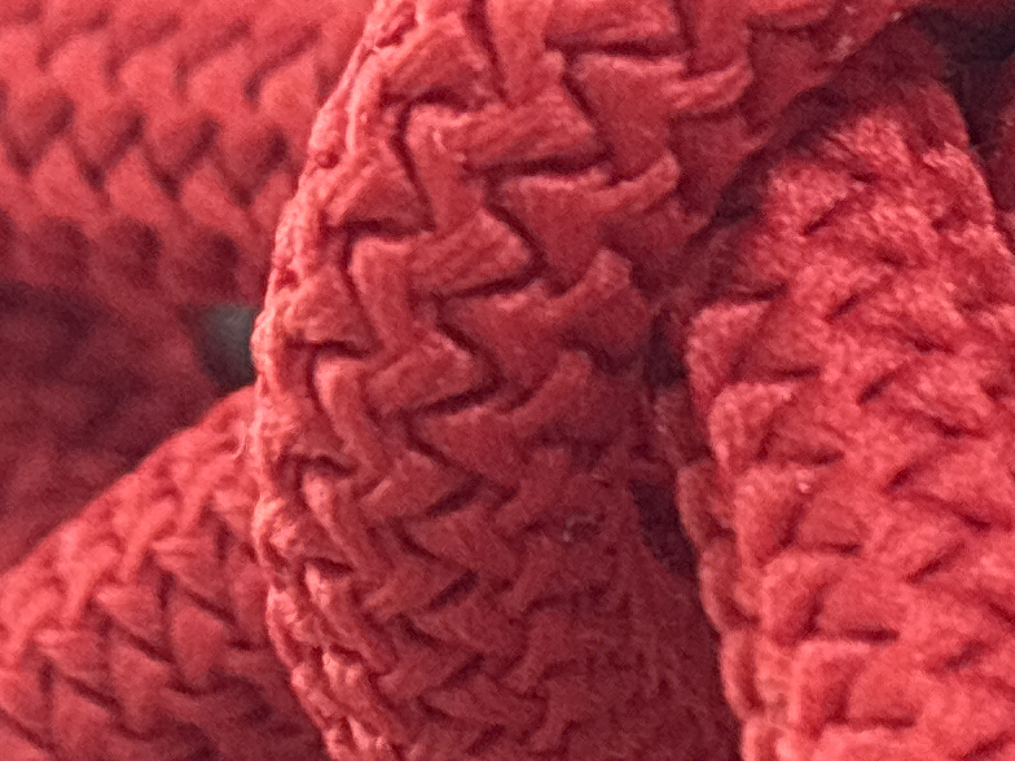 The image displays a close-up, abstract view of a textured, red knitted material. The pattern appears to be a chevron or zig-zag stitch, with thick yarn creating raised ridges and recessed channels. The lighting creates shadows that emphasize the depth and texture of the knit. There are no people, animals, or identifiable objects in the scene. The image does not provide any visual cues or context to associate it with the city of Zvenigorod, Russia, or any specific location.