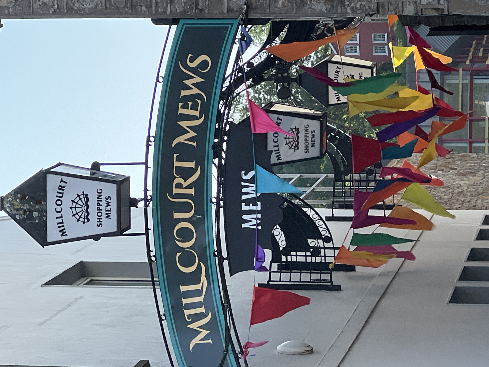The image captures an outdoor scene in what appears to be a pedestrian alleyway or street in Donegal, Ireland. A large, dark green sign with gold lettering arches over the pathway, reading "MILLCOURT MEWS." Below this, another black sign reads "MEWS." Two ornate street lamps are visible, each featuring a white panel with black lettering. The left lamp reads "MILLCOURT SHOPPING MEWS" and has a stylized drawing of a fan-like object over waves. The right lamp is partially obscured but appears to have similar text. A string of colorful triangular flags (bunting) in shades of red, orange, yellow, green, blue, and purple is strung across the pathway, adding a festive touch. The buildings on either side are made of stone and white render. In the background, more foliage and buildings are visible, including a red building with white-trimmed windows. The lighting suggests it is daytime, with sunlight casting shadows and highlighting the colors of the flags. No people are present in the immediate scene.