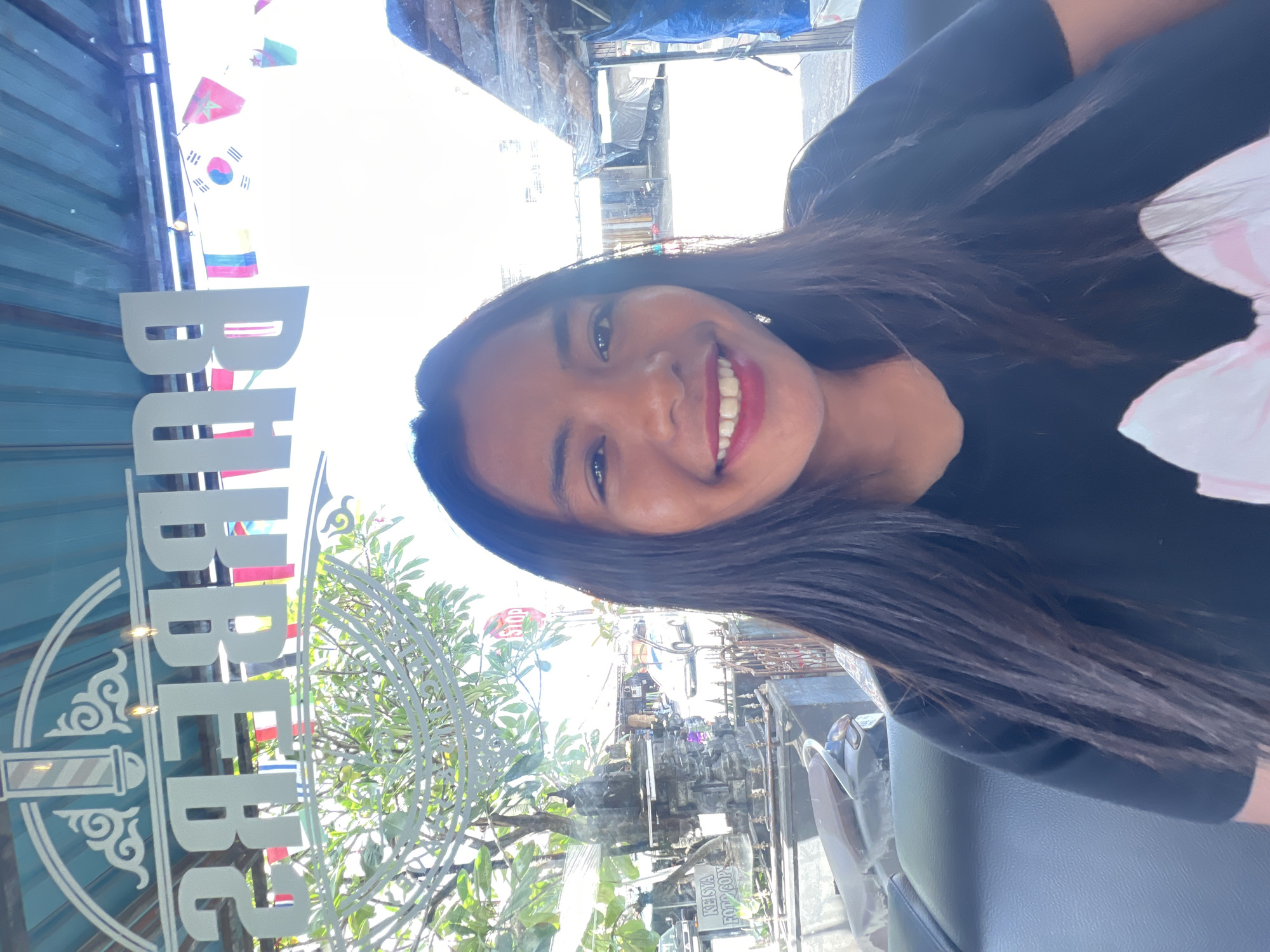 A young woman with long dark hair, wearing a black t-shirt with a large pink bow graphic, smiles at the camera. She is seated inside what appears to be a barbershop, reflected in the glass window behind her. The reflection shows the words "BARBER SHOP" in large, white, reversed lettering across the top. String flags of various countries, including South Korea and Morocco, are visible hanging outside the shop. Through the window, a street scene in Denpasar, Indonesia is visible, with a stop sign, a stone monument, a scooter, and a white car on the road. The environment appears to be a sunny, outdoor commercial area.