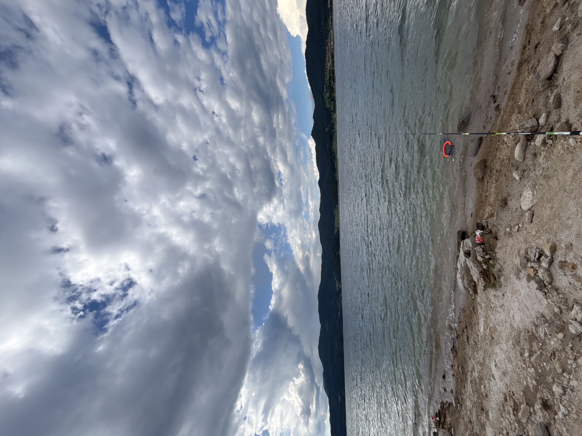 An outdoor, eye-level, medium shot captures a scenic view of a lake under a dramatic sky. The foreground is dominated by a rocky, sandy shore with scattered stones and some visible debris, including a red and white plastic bag. A fishing rod stands planted in the sand near the water's edge, with a small, red, circular flotation device bobbing in the shallow water. The lake itself stretches across the middle ground, its surface rippled by a gentle breeze. In the distance, a range of dark, tree-covered hills forms the horizon, with hints of buildings visible on their slopes. The sky above is a dynamic spectacle of clouds, with patches of bright blue sky peeking through dramatic formations of dark, heavy clouds, suggesting an impending storm or recent rain. The overall impression is one of natural beauty, with a solitary fishing rod hinting at human presence in an otherwise wild landscape.