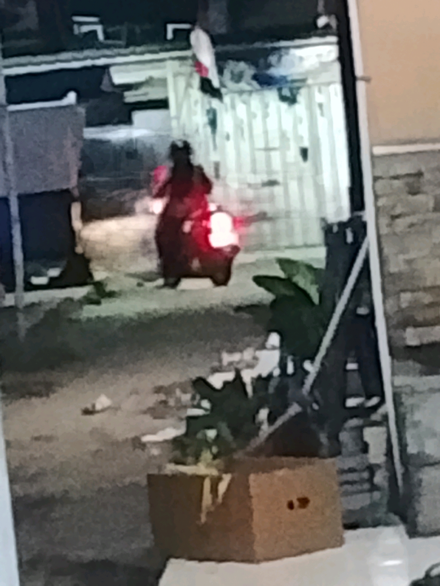 A person wearing a red garment is riding a motorcycle at night in what appears to be a narrow alleyway in Ujungrusi, Indonesia. The motorcycle's headlight and taillight are illuminated, casting a glow on the ground. The surrounding environment is dimly lit, with buildings or structures visible on either side of the alley. A cardboard box is present in the foreground, and there are some plants to the right of the frame. The image quality is low, with significant blur and noise, making it difficult to discern fine details.