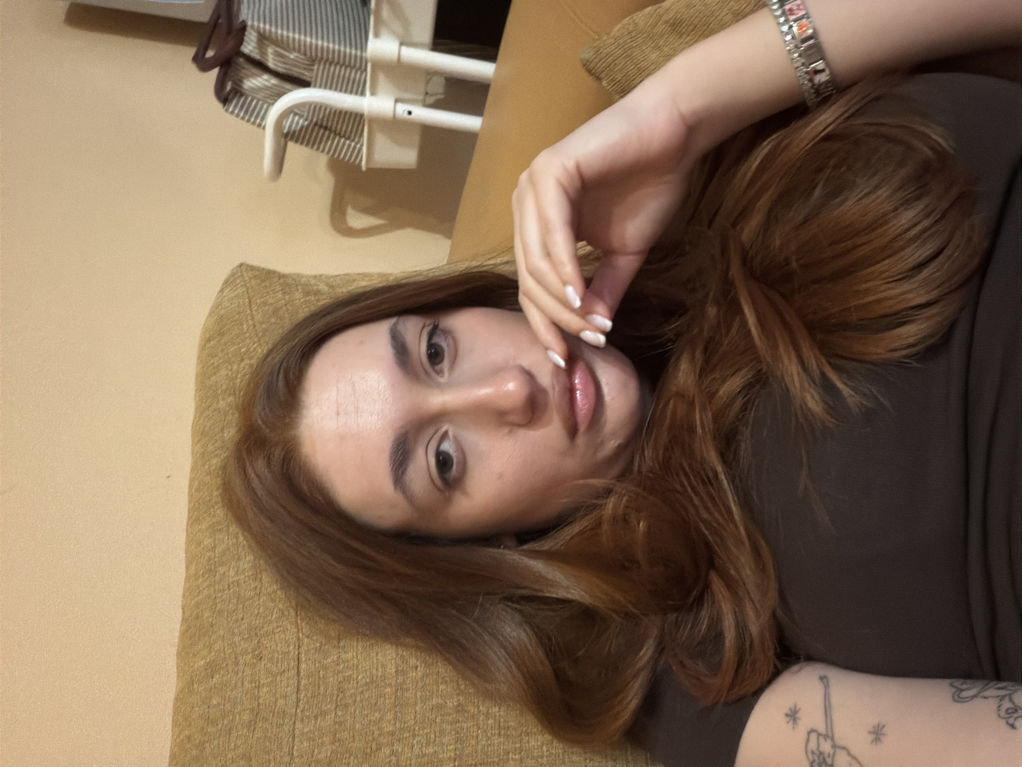 A woman lies on a brown couch. She has fair skin, brown hair, and light-colored fingernails. She wears a bracelet on her left wrist and has tattoos on her left forearm. The background includes a cream-colored wall, a striped bag, and a white metal frame. The location is not apparent from the provided image.
