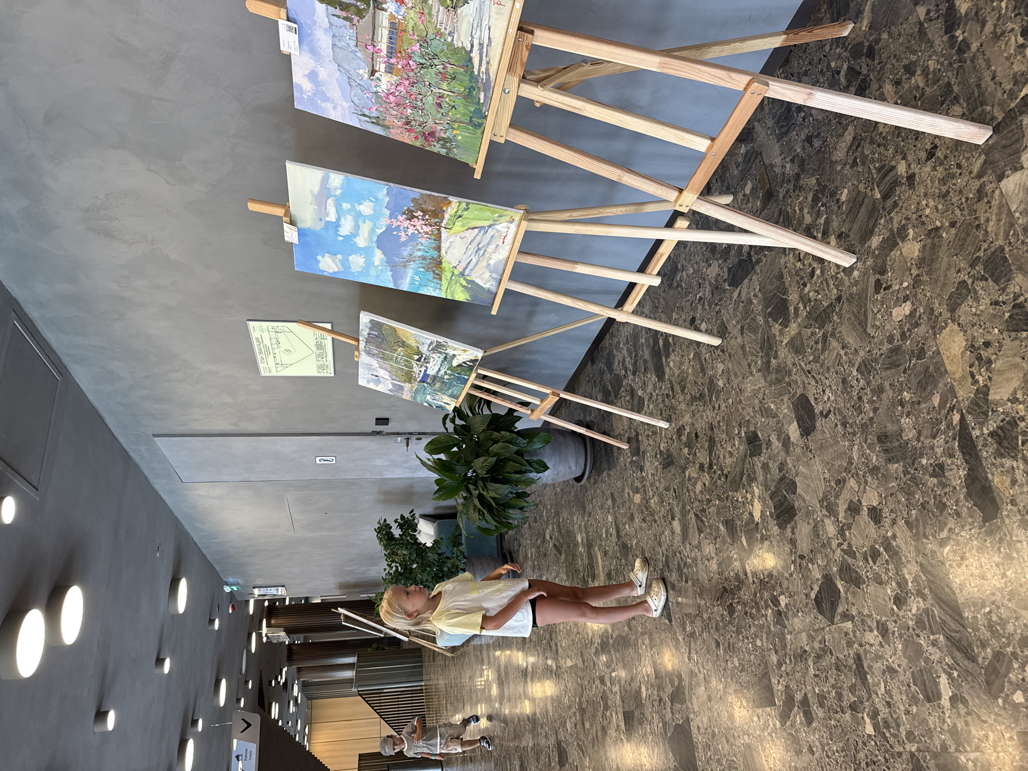 A young girl with blonde hair stands in a modern building hallway, facing a display of paintings on easels. She is wearing a yellow t-shirt and black shorts, and white clogs. The paintings depict landscapes and seascapes. In the background, a young boy is walking. The hallway has a gray, textured wall and a dark, speckled floor. There are also potted plants and overhead lighting. The overall impression is of an art exhibition or a public space with art displayed.