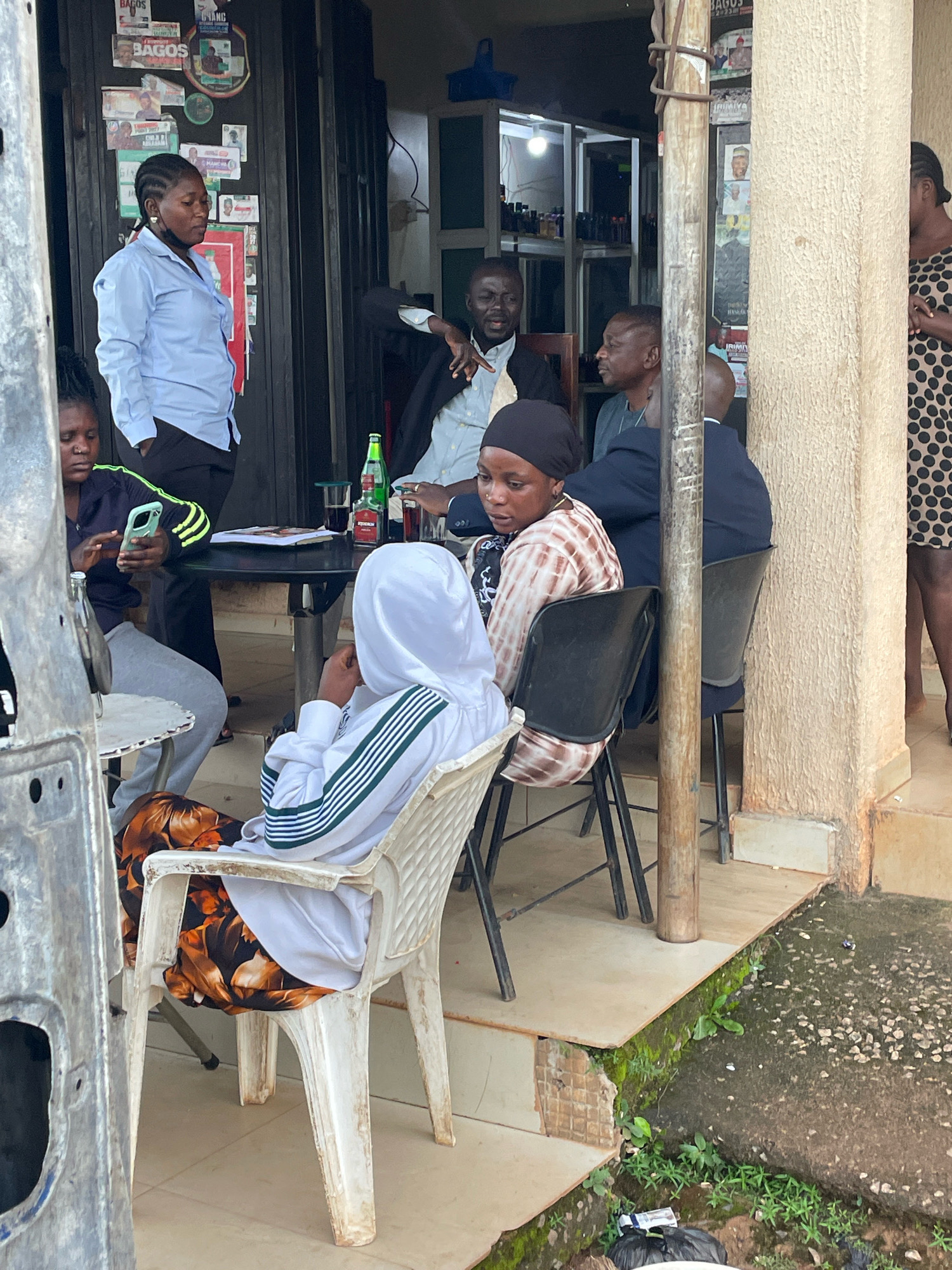 The image depicts an outdoor scene in Jos, Nigeria. In the foreground, a person sits in a white plastic chair, wearing a white hooded jacket with green and white stripes on the sleeves and a patterned skirt. To their left, another person is seated in a similar chair, looking at a smartphone and wearing a dark blue top with yellow stripes.
Further back, a group of people are gathered around a black circular table. One man in a light blue shirt stands near a doorway, while another man in a dark jacket and white shirt is seated. A third man in a dark blue suit is also seated at the table. On the table are drinks and papers.
The environment appears to be a casual outdoor seating area, possibly a roadside establishment. Posters are visible on a dark door or wall behind the standing woman. A pillar, likely part of a building structure, is prominent in the right side of the image, with a rough, textured surface. The ground is paved with tiles in some areas and a rough, stony surface in others, with some greenery growing in cracks and along the edges. Some debris, including a black trash bag, is present on the ground. A portion of a burnt vehicle, with visible metal and wires, is present in the far left foreground.