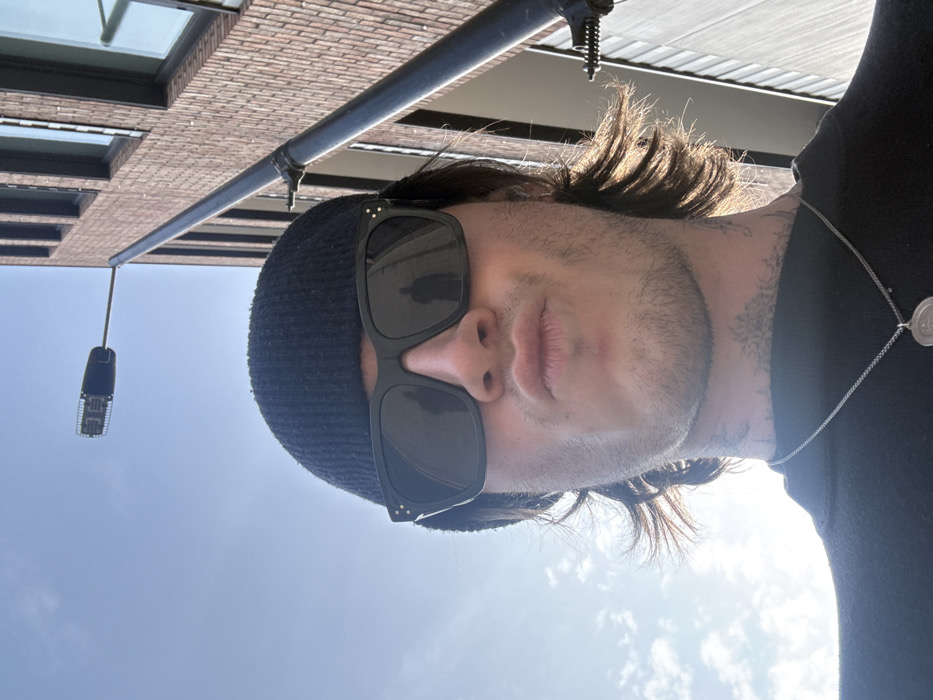 An upward-angled selfie captures a male subject in an urban outdoor environment, identified as the City of London, United Kingdom. The subject, positioned centrally, wears a black knitted beanie, black square-framed sunglasses, and a black t-shirt with a silver chain necklace featuring a circular pendant. He has stubble on his chin and jawline, and medium-length dark hair is visible beneath the beanie, back-lit by strong sunlight. Tattoos are visible on the side of his neck. His lips are pursed in a "pouting" expression. Reflections in the sunglasses show two dark, human-like silhouettes.
The background consists of a clear blue sky with minimal cloud cover, indicating bright daylight. To the right, a tall black pole supports a modern LED street light fixture. Behind the pole and further right, a multi-story building constructed of dark reddish-brown bricks with dark-framed windows is partially visible, consistent with contemporary urban architecture.
