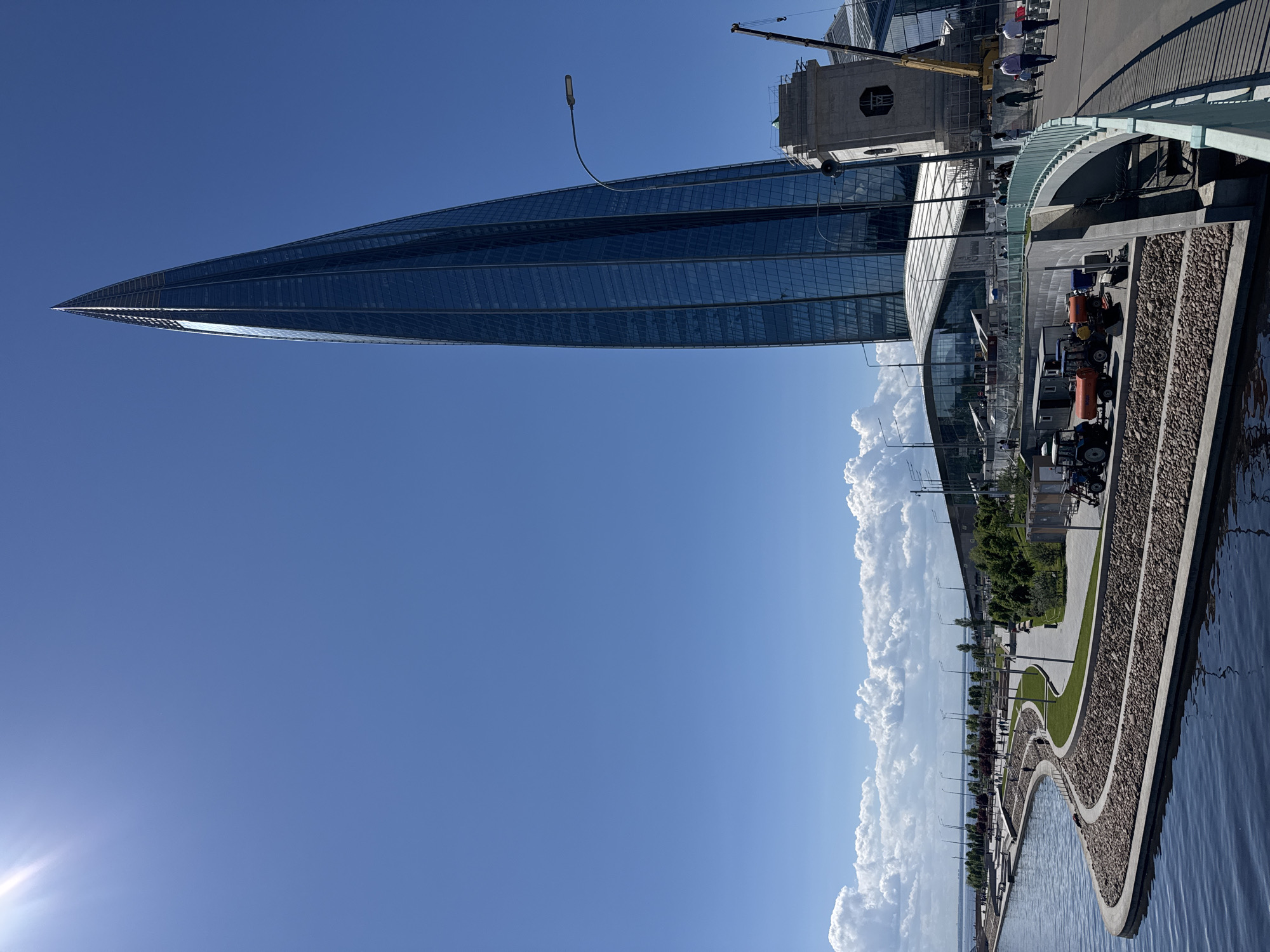 The image captures a modern architectural landscape on a bright, sunny day in Saint Petersburg, Russia. The most prominent subject is the Lakhta Center skyscraper, a towering, slender structure clad in reflective glass that tapers to a sharp point, dominating the left side of the frame and reflecting the expansive blue sky. Adjacent to it, a lower, curved glass building and a concrete structure, partially obscured by a yellow construction crane, are visible, hinting at ongoing development or maintenance.
In the foreground and extending to the right is a wide body of water, likely the Gulf of Finland or an inlet. A modern, winding promenade runs along the waterfront, featuring light-colored paved paths, manicured green lawns, and sections of decorative gravel. Contemporary streetlights are spaced along the promenade, which is also lined with small trees and bushes.
Several utility vehicles are parked on the embankment, including orange tractors with "AVRORA 1500" visible on their sides, and a green mini-tractor. A few people are seen walking casually on the promenade and near the buildings, appearing small in scale compared to the massive architecture.
The time of day is bright daylight, with strong sunlight creating clear shadows and a lens flare visible in the bottom left, suggesting either morning or late afternoon. The sky is a vibrant, clear blue with a line of fluffy white cumulus clouds along the distant horizon. The scene depicts a clean, well-maintained, and modern urban environment under pleasant weather conditions.