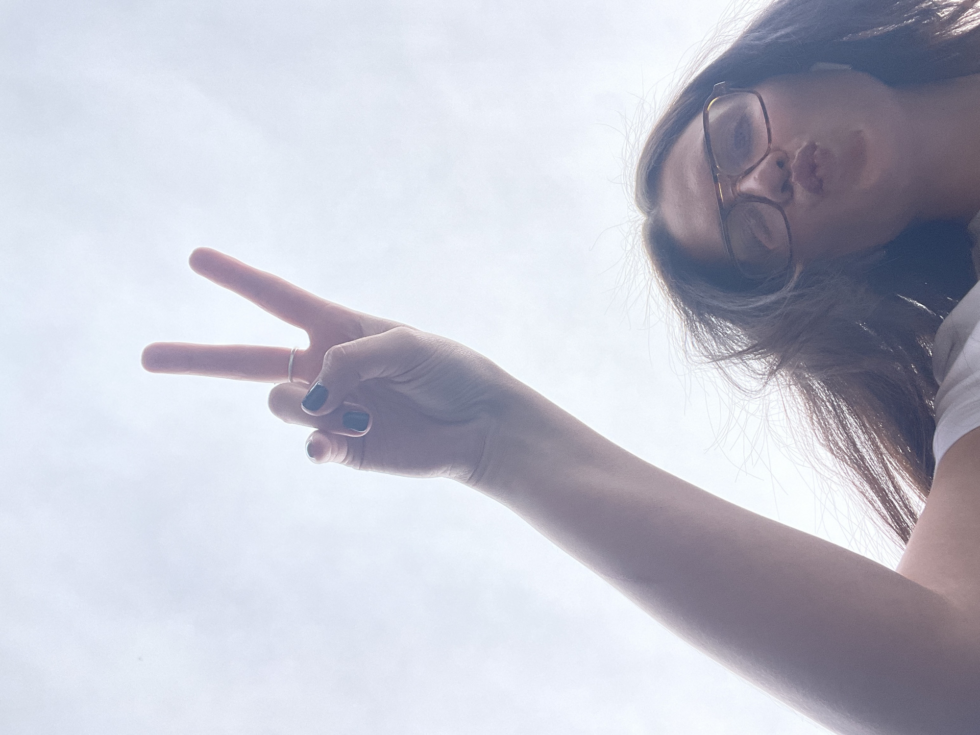 A low-angle shot depicts a woman with long brown hair and glasses making a peace sign with her right hand. She has dark nail polish. Her lips are pursed slightly. The background is a bright, overcast sky. There's no information to deduce any location in Moscow, Russia.