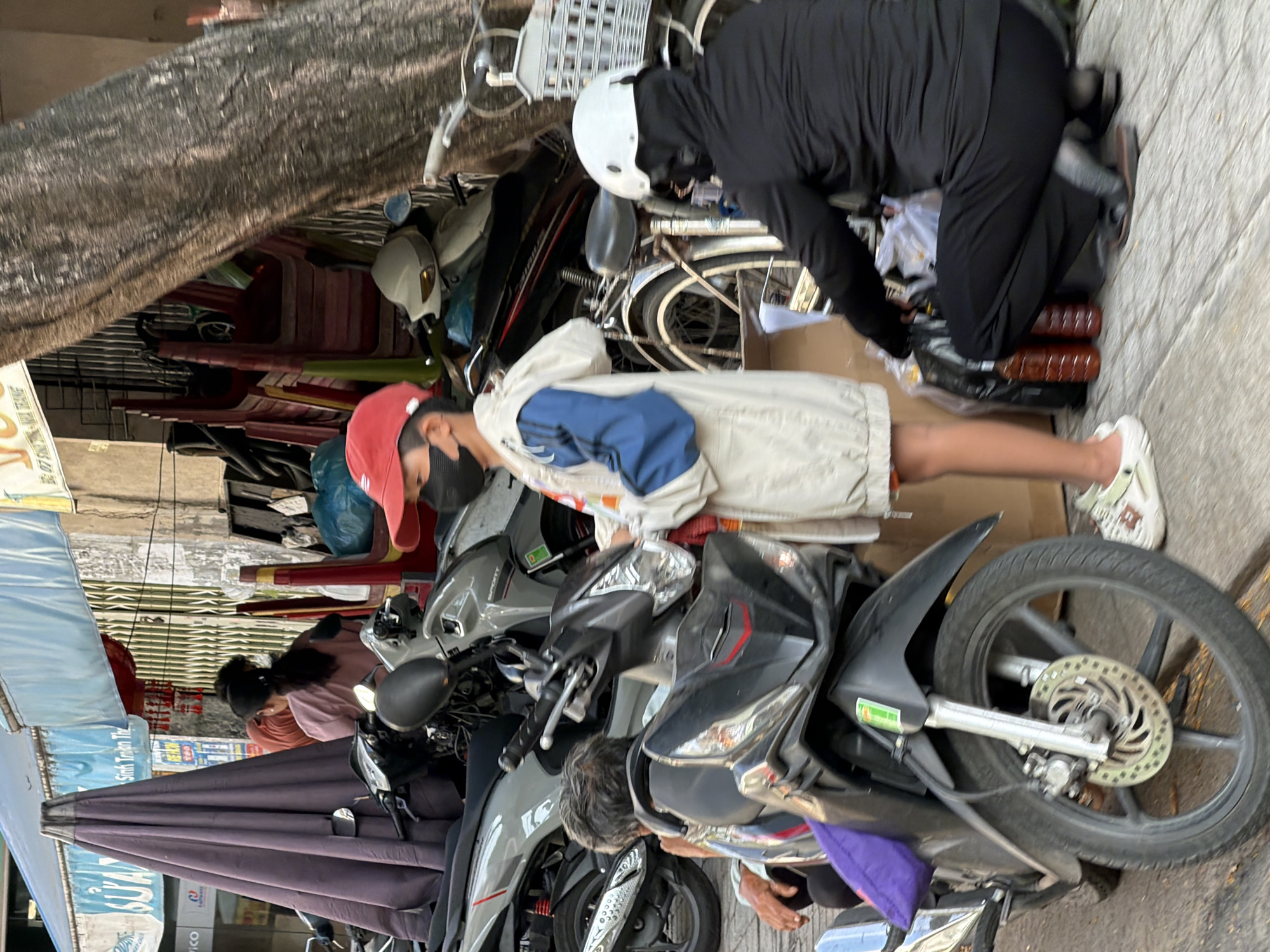 The image captures a street scene in Nha Trang, Vietnam, with several motorcycles parked along a sidewalk. In the foreground, a young person wearing a red cap, a dark face mask, and a light-colored jacket with blue sleeves stands near a parked black motorcycle. To the right, another individual, wearing a black hooded jacket, dark pants, and a white helmet with a black face mask, is crouched down, attending to items on the ground. Several bottles and plastic bags are visible near the crouching person. In the background, more motorcycles, a large purple umbrella, and stacks of chairs suggest a commercial or public area. The overall environment appears to be a busy urban street.