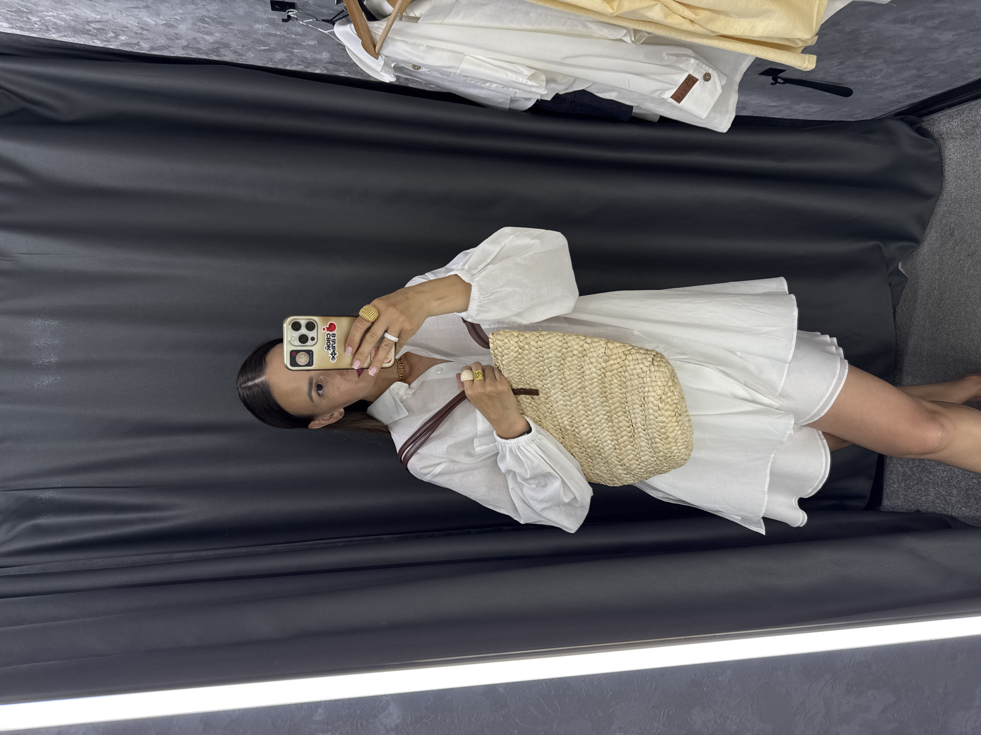 A woman stands in a fitting room, taking a selfie with her phone. She is wearing a white linen dress and holding a straw tote bag. The fitting room has dark gray curtains and a textured wall. Clothes hang on a rack to her right. The image does not provide any information about the city of Sochi, Russia.