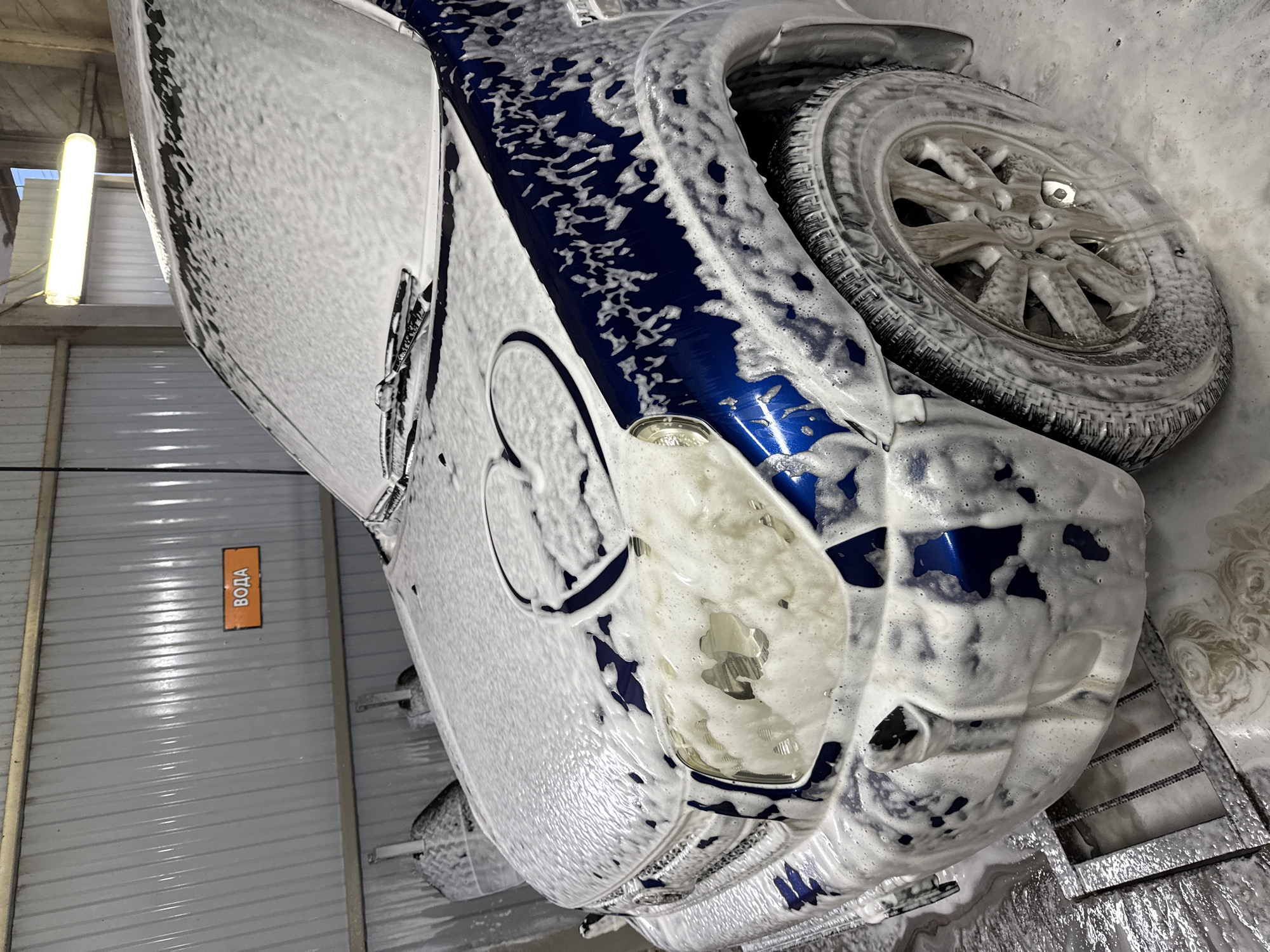 A blue SUV, likely a Toyota RAV4, is depicted within a car wash bay in Chaikovsky, Russia, completely covered in white cleaning foam. A distinct heart shape has been wiped or drawn into the foam on the front left side of the hood, exposing the vehicle's dark blue paint beneath. The front left headlight, part of the grille, windshield with wipers, and the front left wheel with a multi-spoke alloy rim and winter tire are visible through the foam. The car wash environment features walls made of light-colored corrugated metal panels. An orange rectangular sign displaying the Cyrillic word "ВОДА" (VODA), meaning "WATER," is mounted on the wall in the upper left background. A single fluorescent tube light fixture hangs from the ceiling, providing illumination. The wet floor in the foreground shows a metal drainage grate. No individuals are visible in the scene.