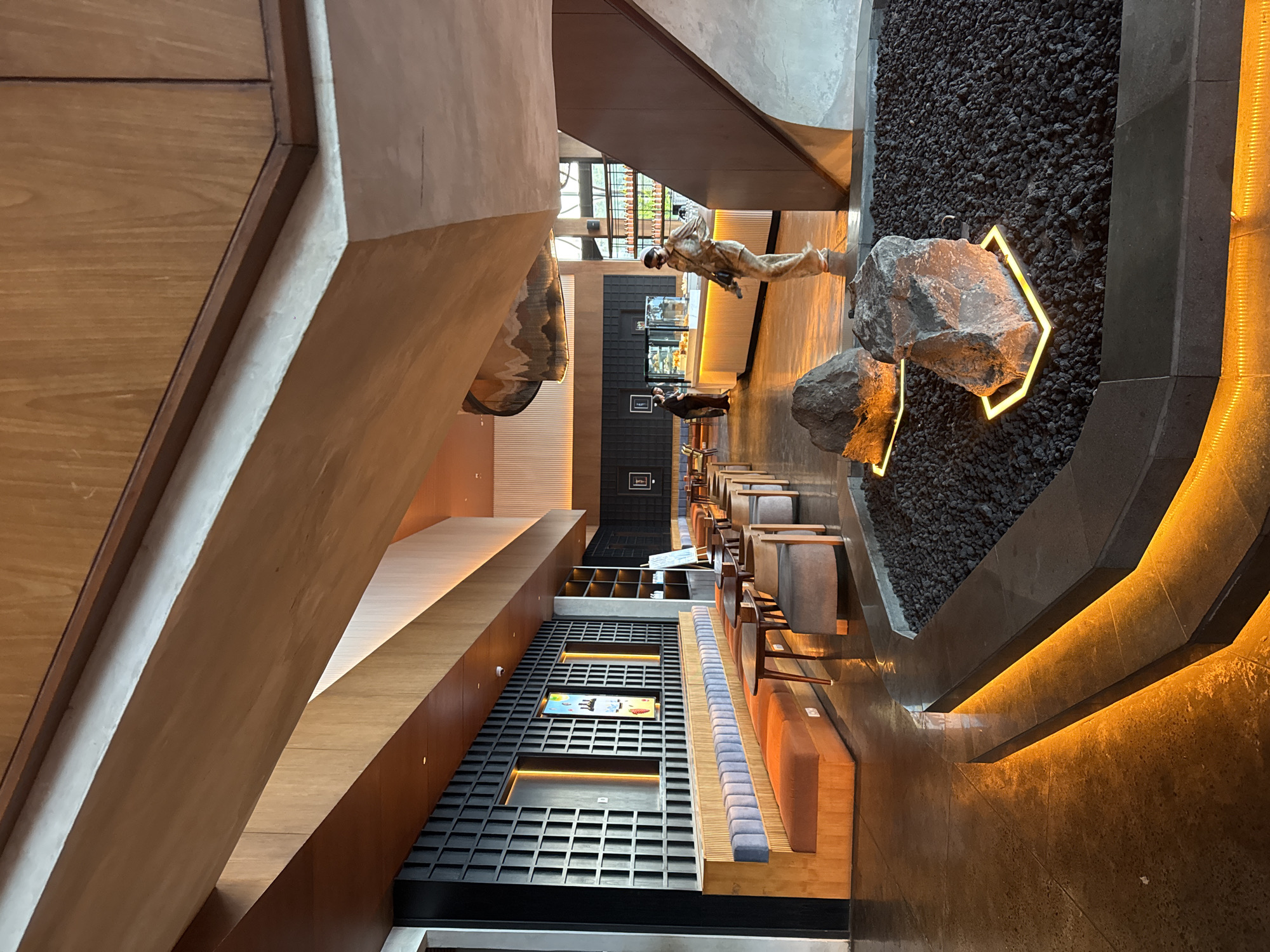 The image shows the interior of a modern, upscale restaurant or cafe in Kintamani, Indonesia, with a focus on natural materials and ambient lighting. In the foreground, two large rocks are placed on a bed of black gravel, each illuminated by a square neon light at its base. The gravel area is bordered by a low, dark stone wall, with bright orange LED lighting embedded along its steps and edges, creating a warm glow on the polished concrete floor.
The restaurant features a long, built-in banquette seating area with orange and light blue upholstery, facing small wooden tables and chairs. Behind the banquette, a black, textured wall with a grid pattern serves as a backdrop, with two framed artworks and two illuminated rectangular panels mounted on it.
Further into the space, a counter area is visible, with glass display cases showing pastries. Several individuals are present: two people are standing at the counter, appearing to be customers or staff, while another person, a woman in a patterned outfit and sunglasses, is walking away from the counter towards the foreground. She is holding a small bag.
The architecture of the space incorporates large wooden beams and panels, with a unique, curved ceiling feature above the counter area adorned with a patterned design. Natural light streams in from windows in the background, which reveal glimpses of foliage, suggesting an outdoor view. The overall ambiance is sophisticated and contemporary, blending natural elements with modern design.