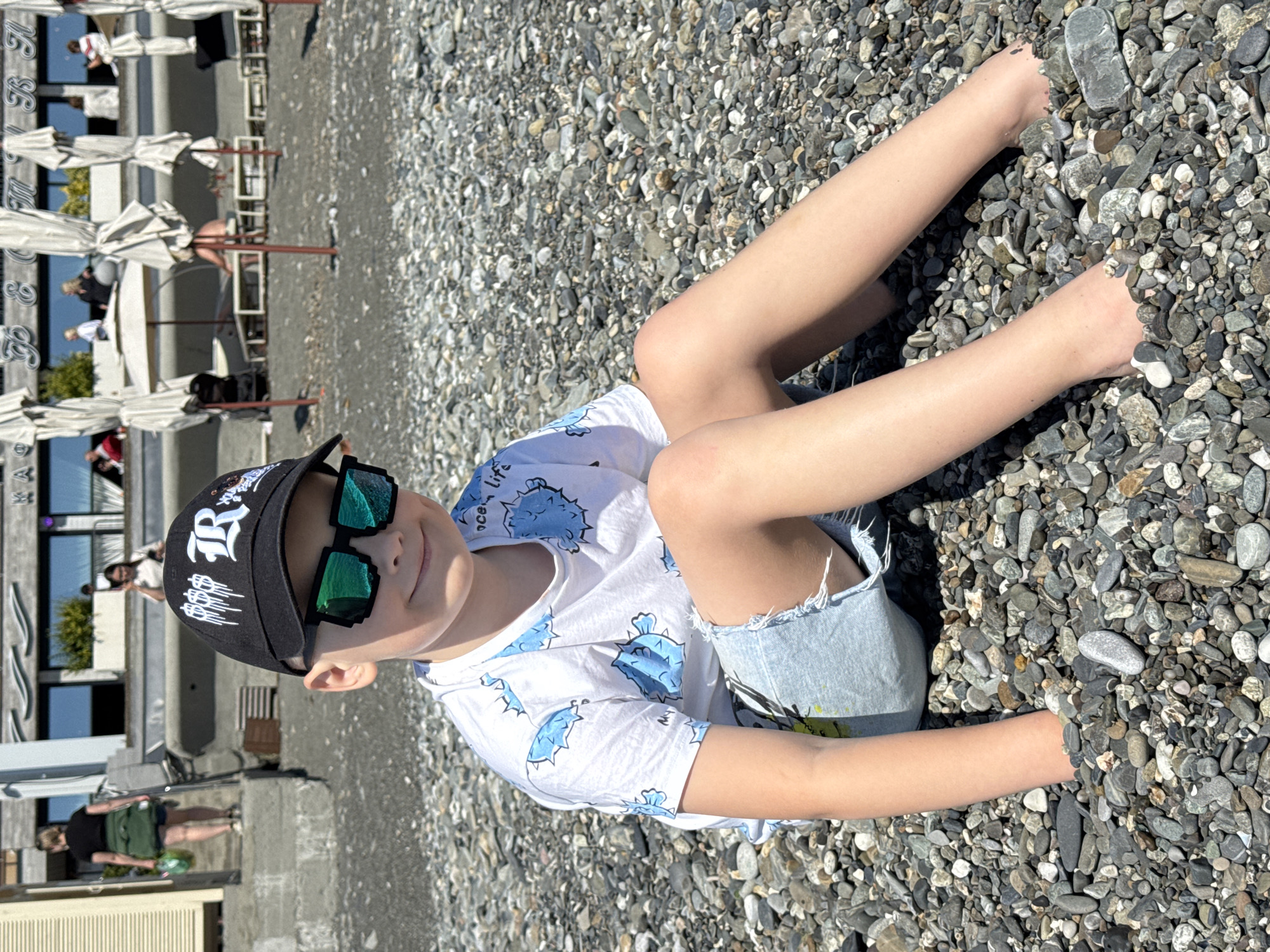 A child wearing a baseball cap and sunglasses sits on a pebble beach, possibly in Sochi, Russia. The child is wearing a white t-shirt and denim shorts. There are buildings and people in the background. Umbrellas and beach chairs can also be seen.