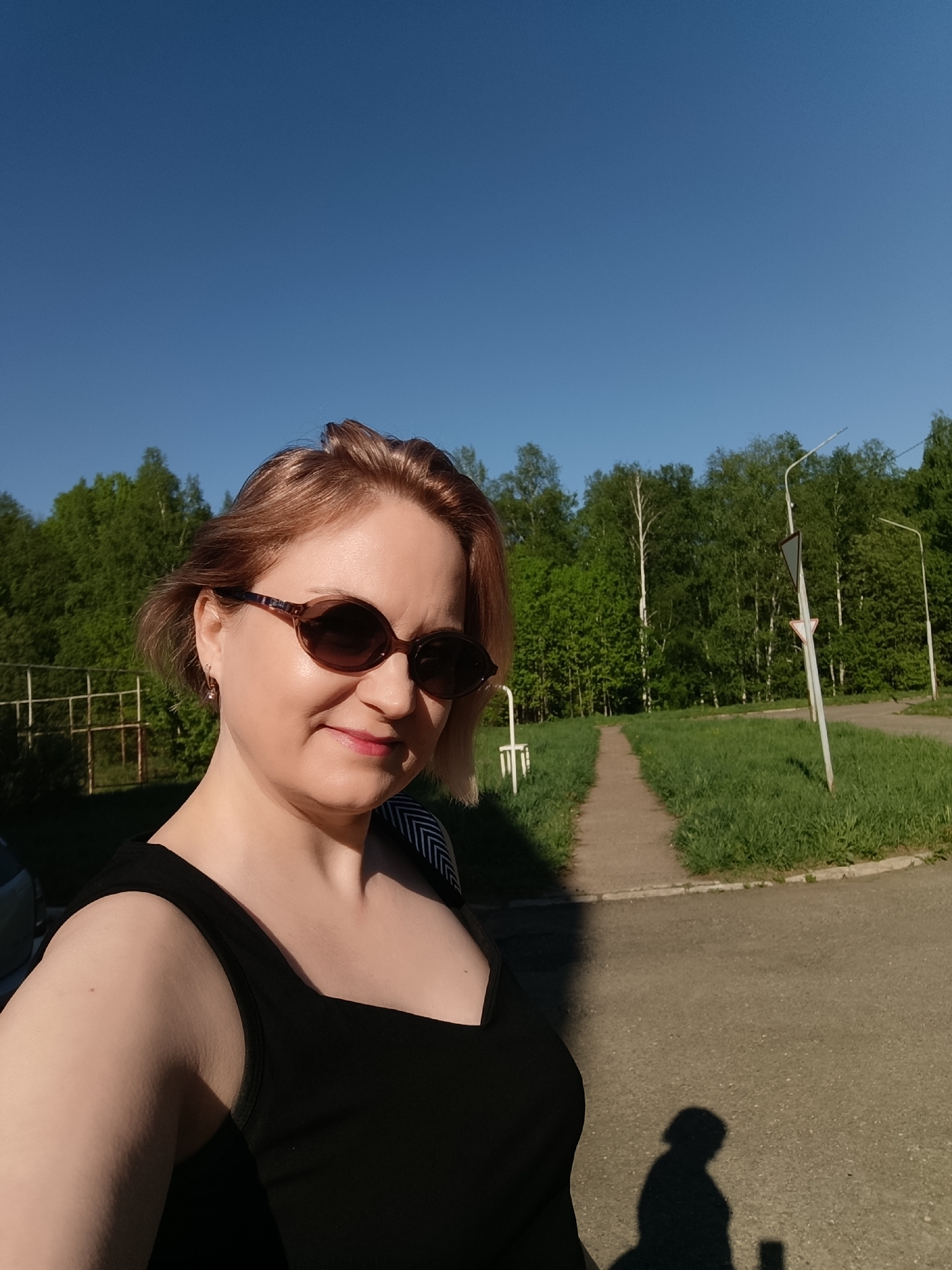 A woman with short, light brown hair and sunglasses stands on a paved area in what appears to be an urban environment, likely in Марковский, Russia. She wears a black tank top. Behind her is a grassy area, a walking path, trees, and streetlights. The sky is blue. Her shadow is visible on the pavement. There is also a fence and another vehicle in the background.