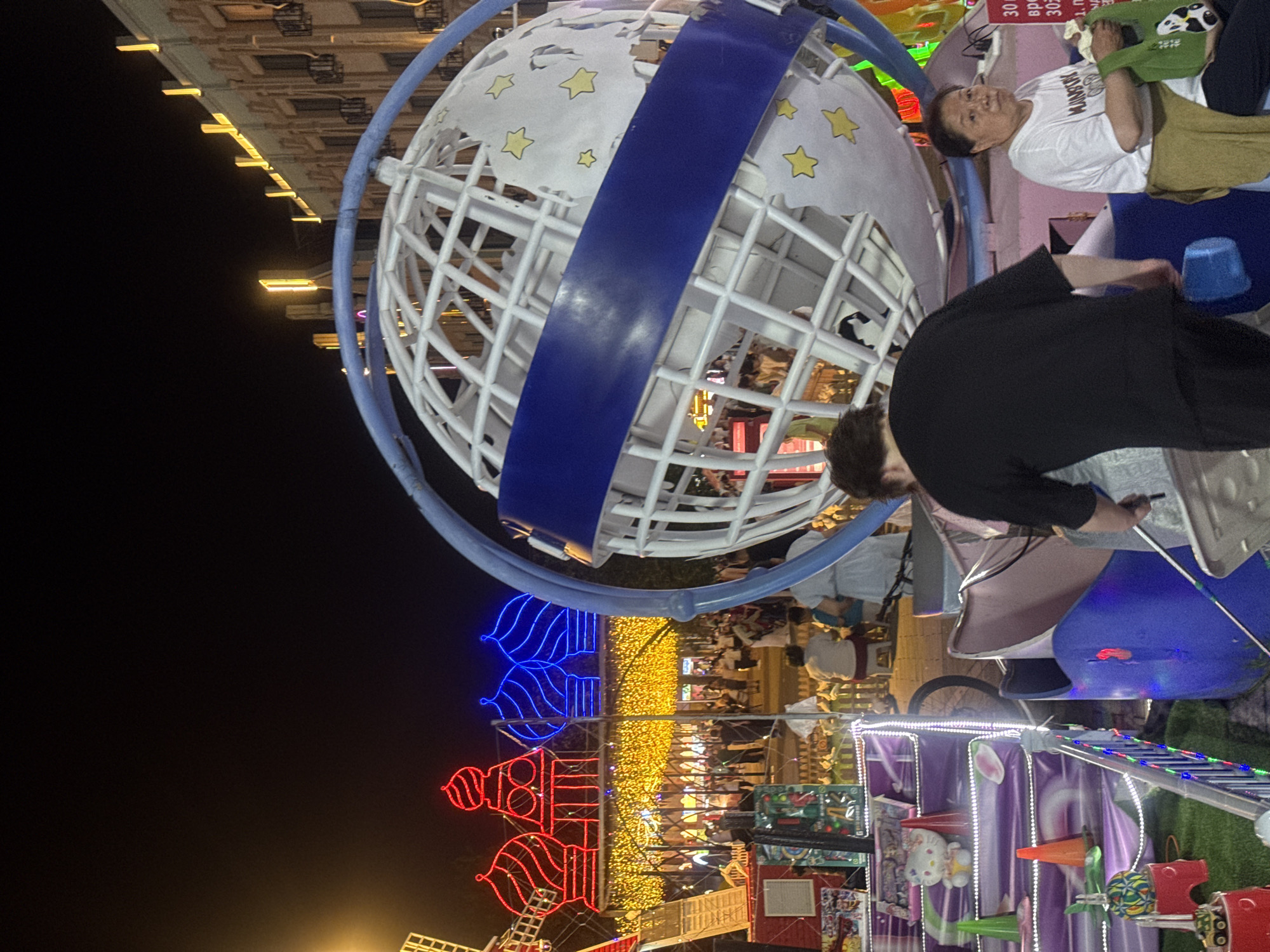 The image depicts a night scene at what appears to be an amusement park or carnival in Qinhuangdao, China. In the foreground, a large, white, skeletal globe structure with a blue band is prominent. To the left, a colorful children's ride with various cartoon characters and lights is visible. A man in a black shirt and a woman in a white t-shirt are present in the mid-ground, near the globe. In the background, illuminated buildings with decorative lights and several people walking can be seen, suggesting a bustling public space. The overall atmosphere is festive and lively, characterized by the bright lights and the presence of families and individuals enjoying the night.